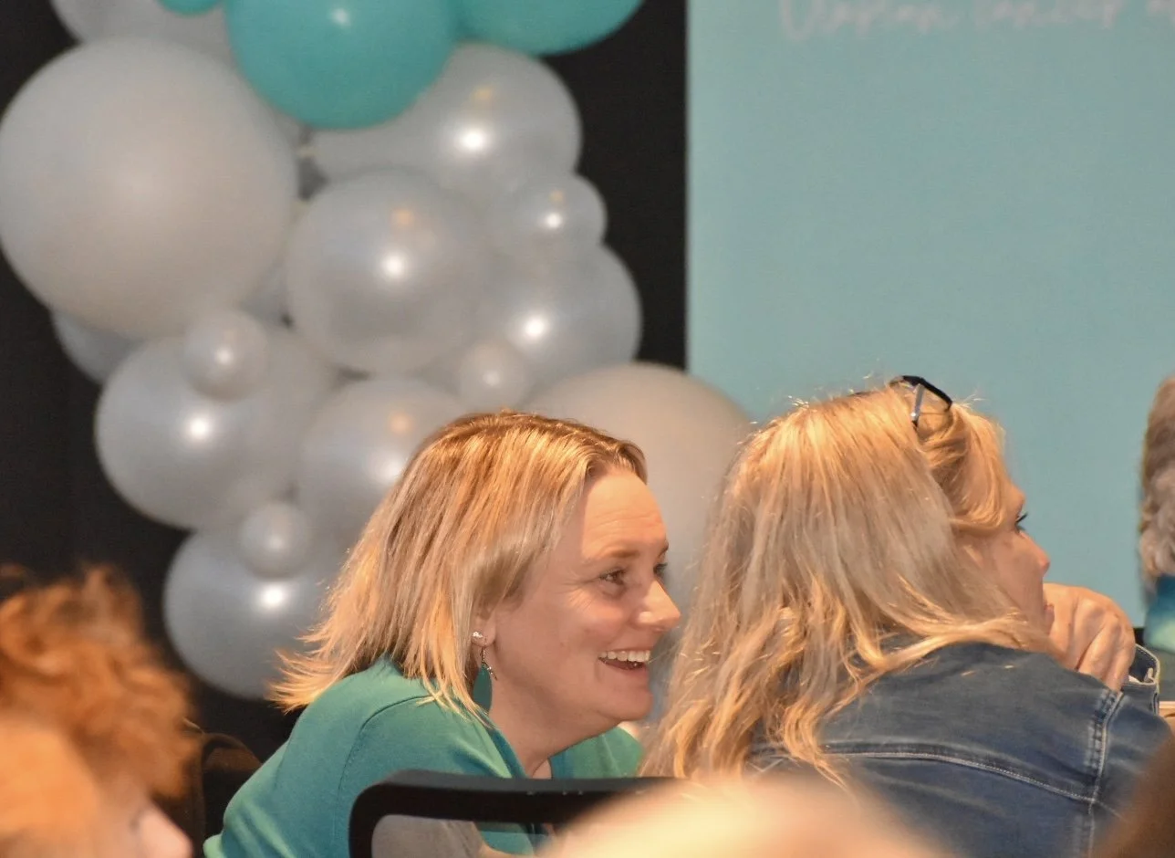 Two women sitting at a table in a room decorated with white and teal balloons, engaged in conversation and smiling.