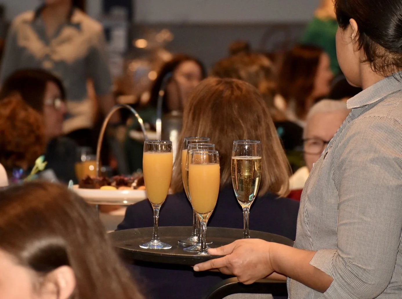 A waitress holds a tray with four glasses of orange and clear beverages at a social gathering or celebration, with many people seated at tables in the background.
