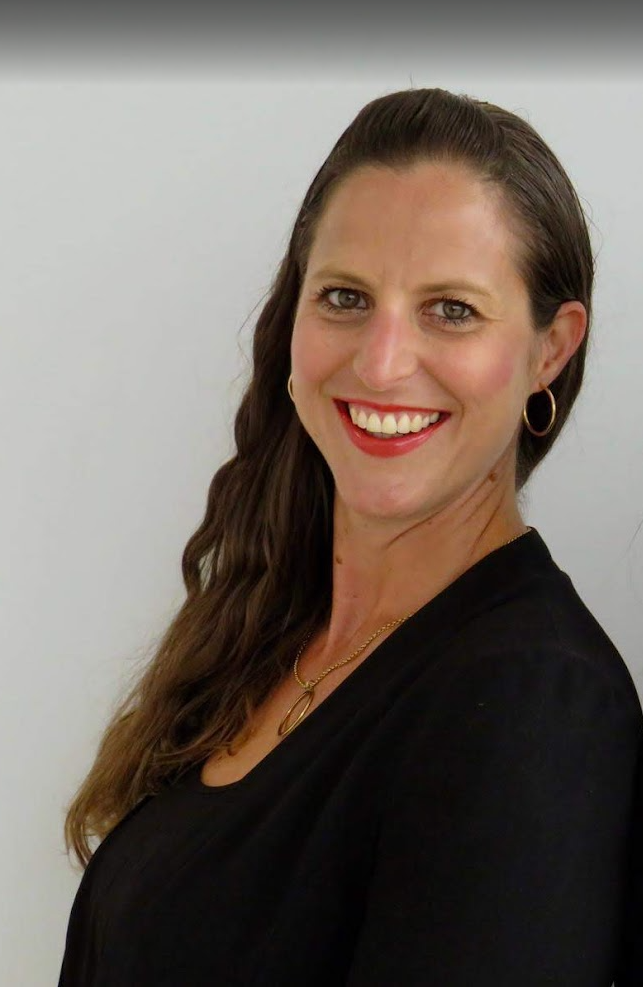 A woman with long wavy brown hair, wearing gold hoop earrings, a gold necklace, and a black top, smiling at the camera against a plain light-colored background.