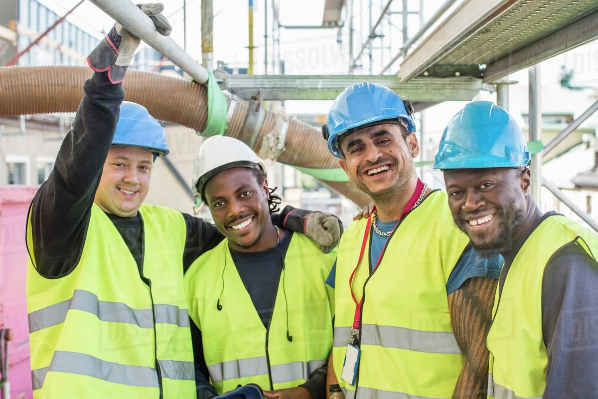 Four construction workers wearing blue and white hard hats and high-visibility yellow vests, smiling at a construction site.
