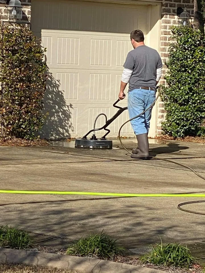 A man in gray T-shirt, blue jeans, and rubber boots is pressure washing a concrete driveway near a garage door, with bushes on either side.