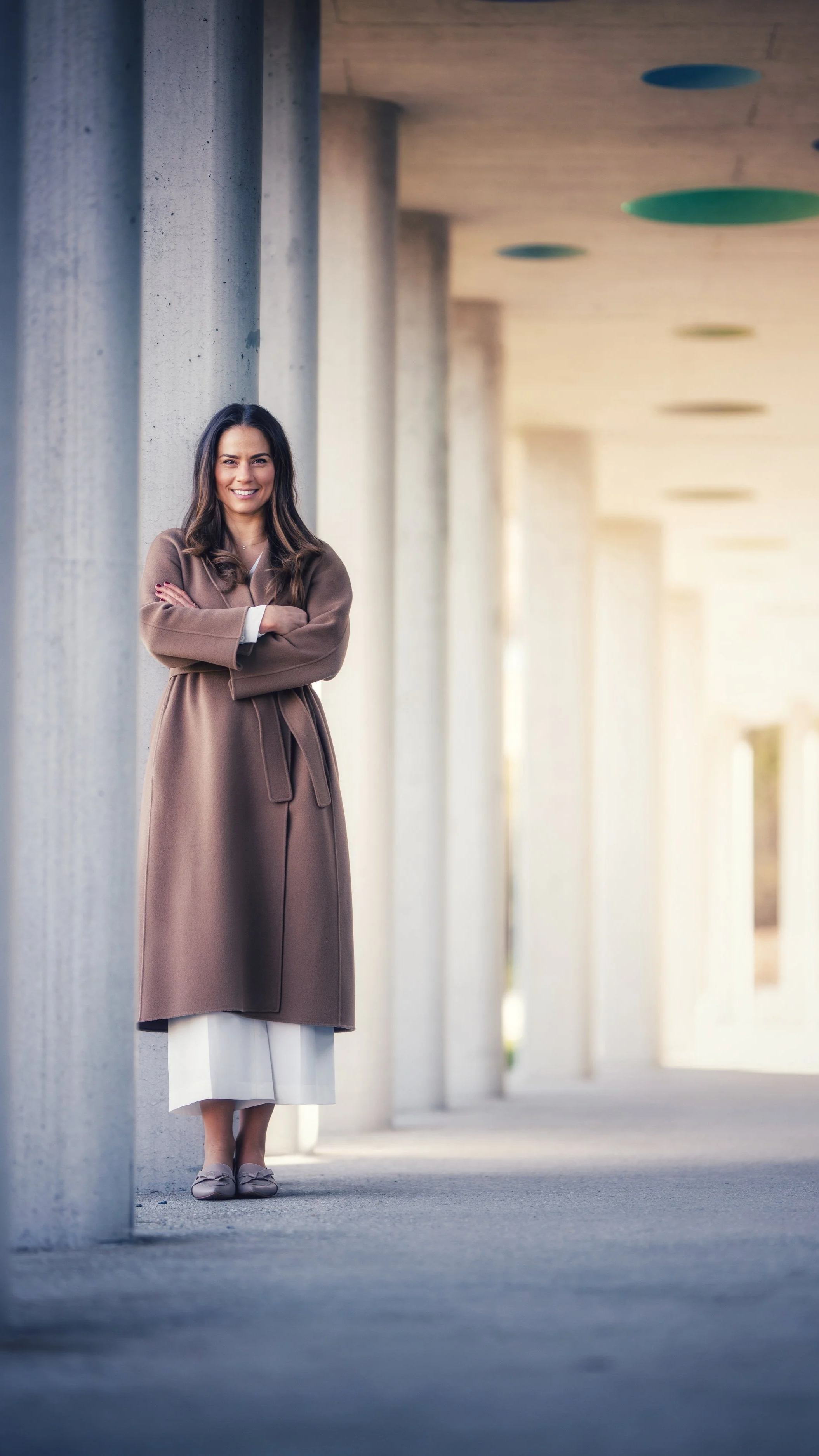 A woman standing outdoors next to large concrete columns, smiling with arms crossed, wearing a brown coat and light-colored dress.