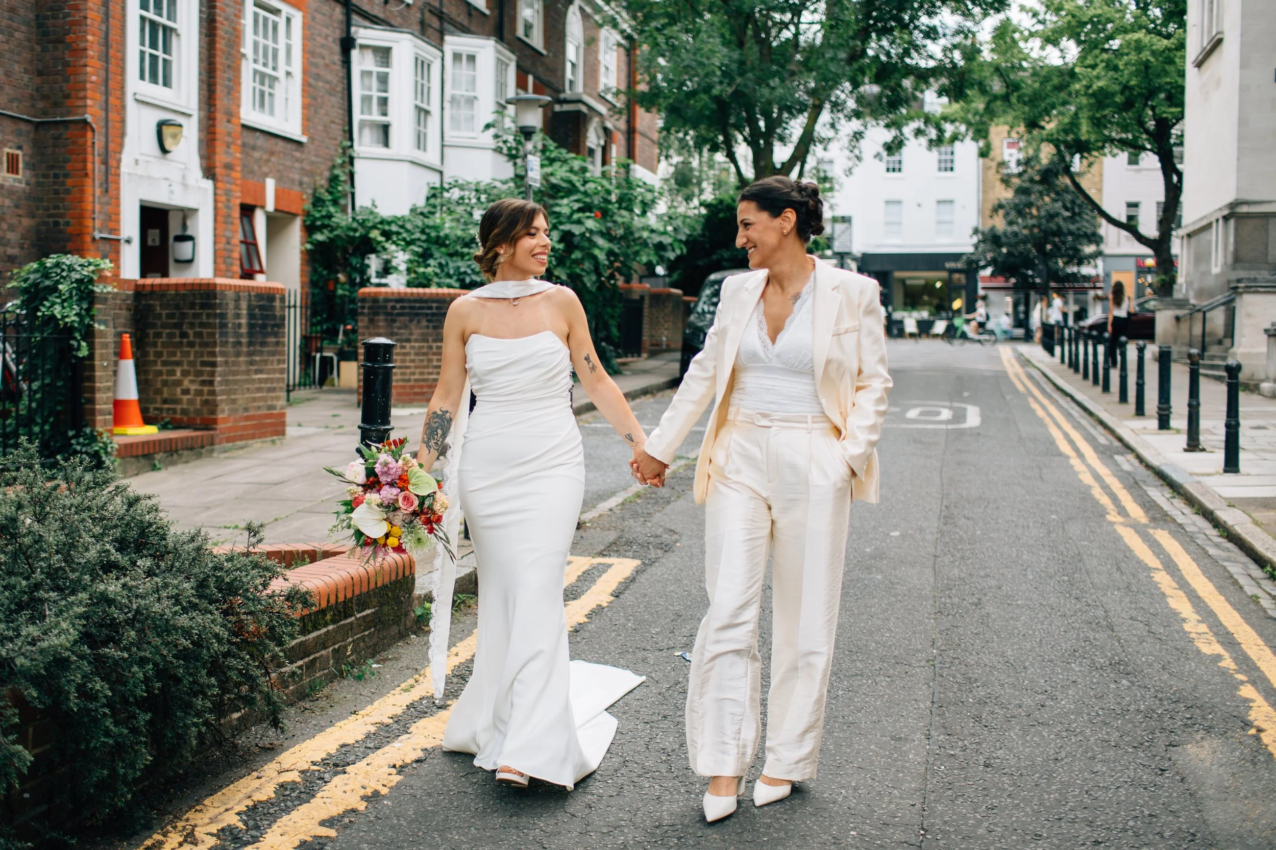 Two women in wedding attire holding hands and smiling at each other on a city street.