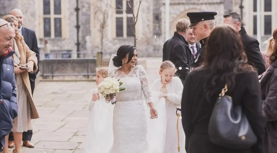 Bride in white wedding dress holding a bouquet, smiling, walking outdoors with children and guests around her on a wedding day.
