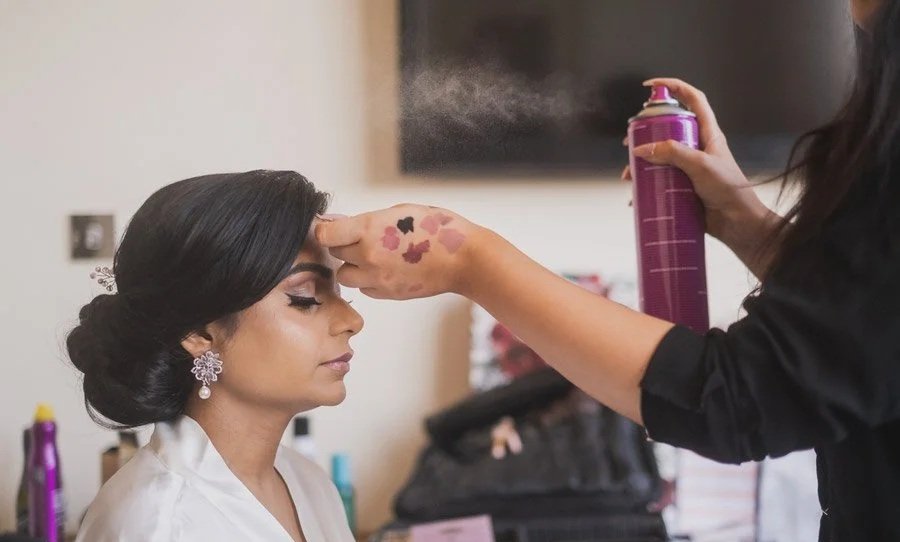 A woman with styled hair and makeup sits calmly with her eyes closed while another person sprays her forehead with hairspray.