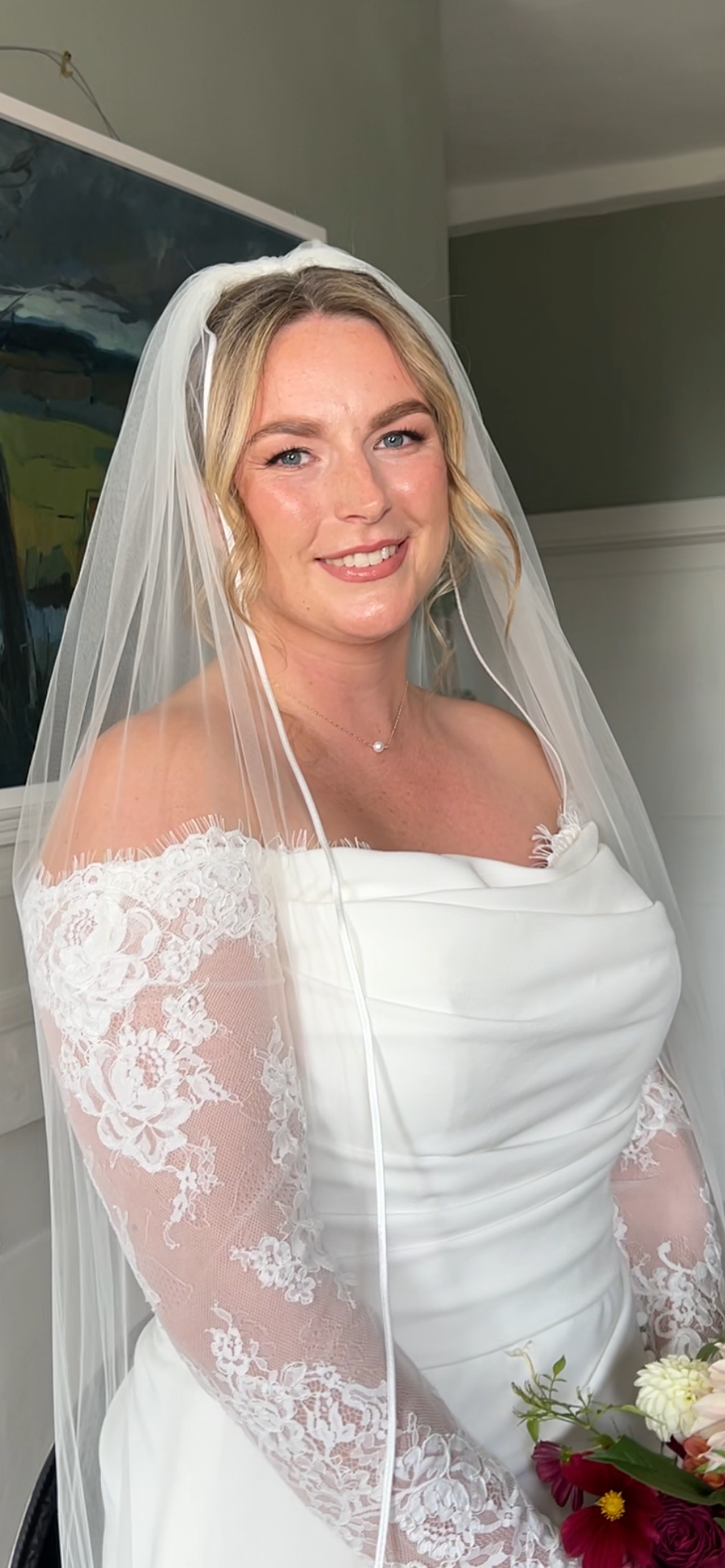 A bride in a white wedding dress with lace sleeves, wearing a veil, holding a bouquet of flowers, smiling indoors.