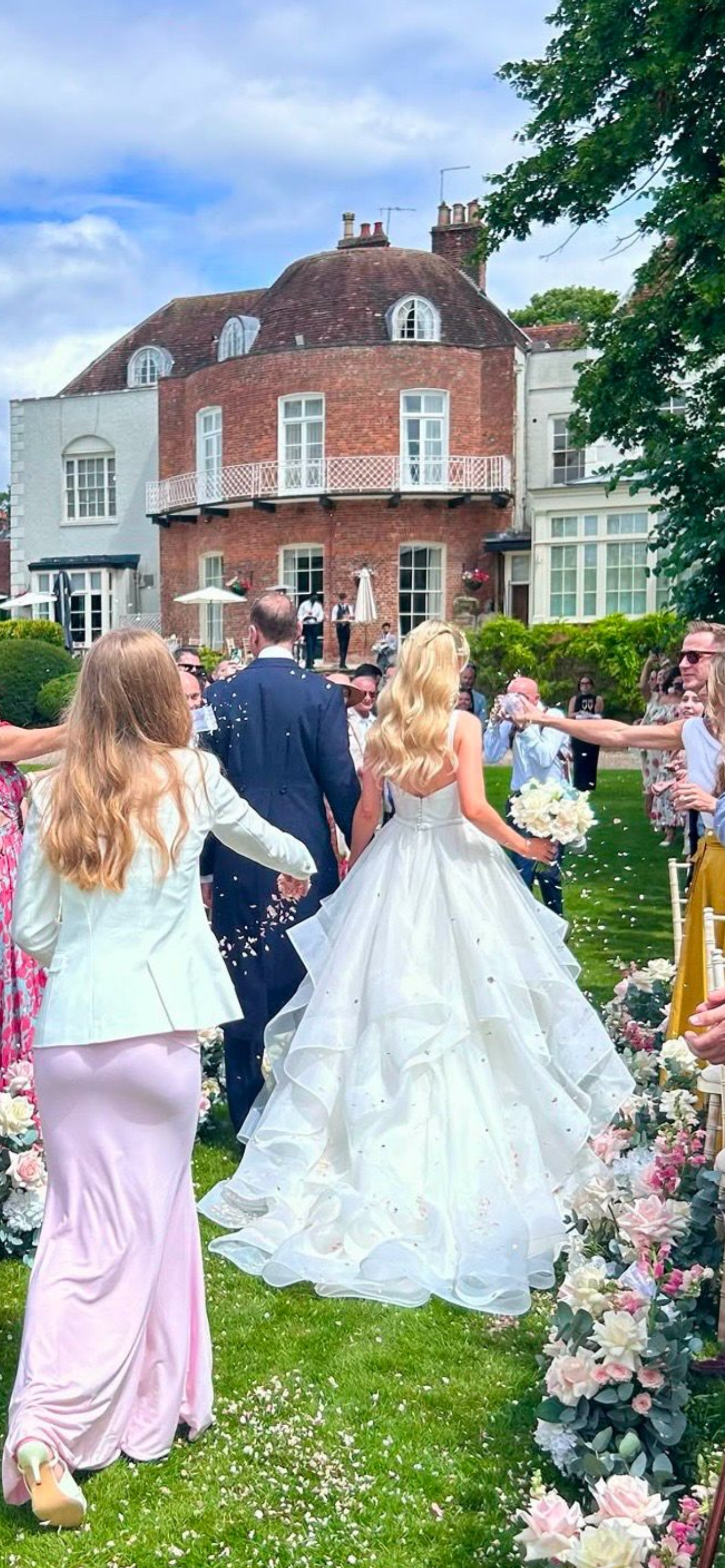 Wedding celebration outdoors with bride in white gown, surrounded by guests, at a garden with a large brick house in the background.