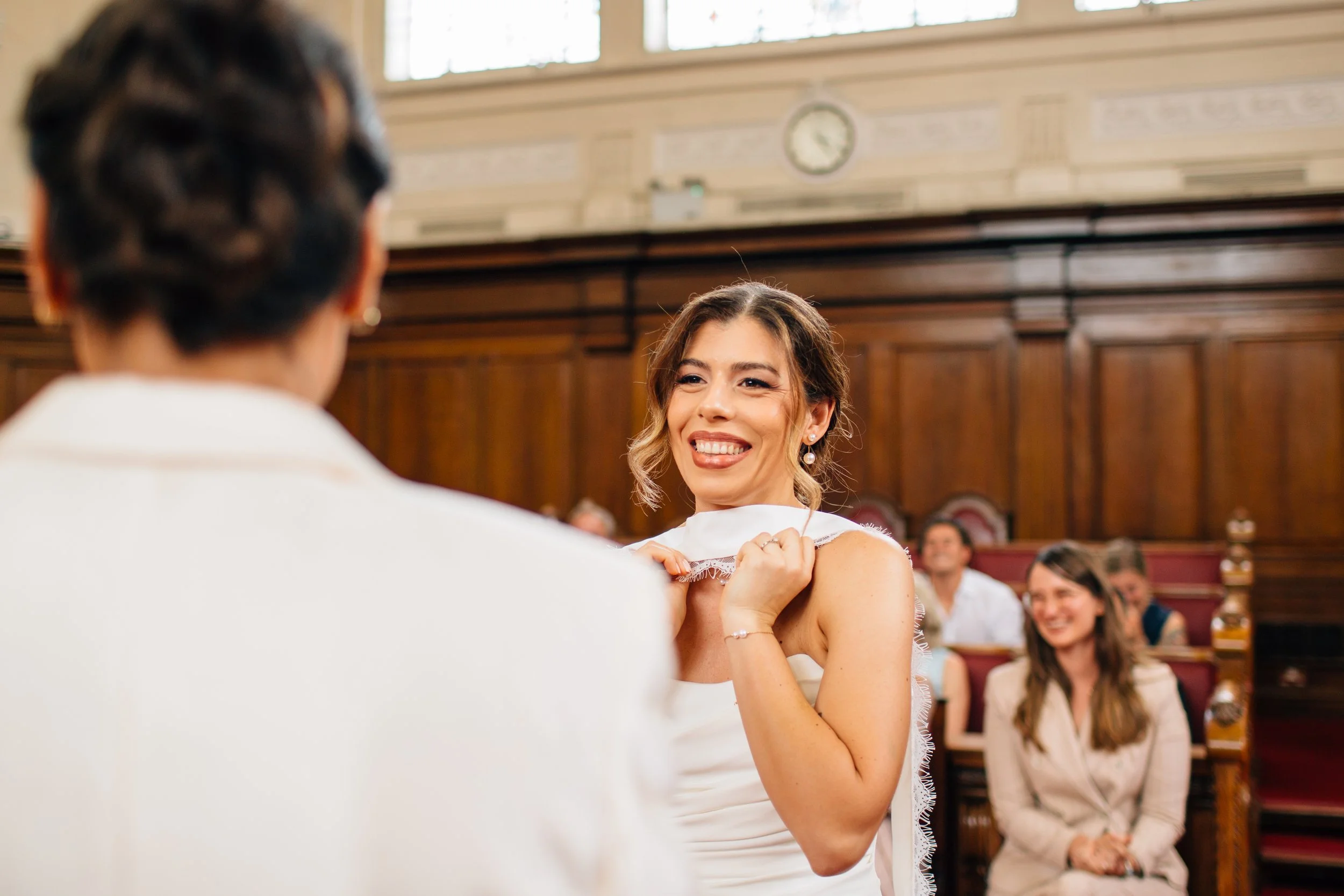 A woman smiling as she is being sworn in during a formal ceremony, with a person standing in front of her and an audience seated in the background.