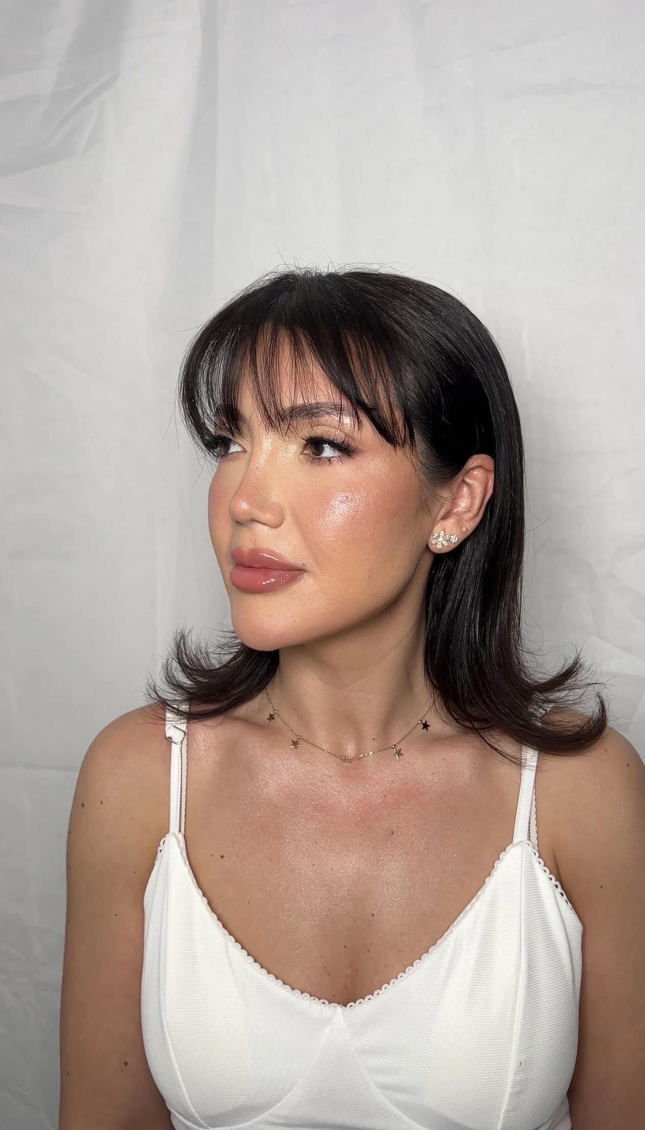 A woman with shoulder-length dark hair and bangs, wearing a white tank top, gold star necklace, and floral earrings, looking to her left against a plain white background.