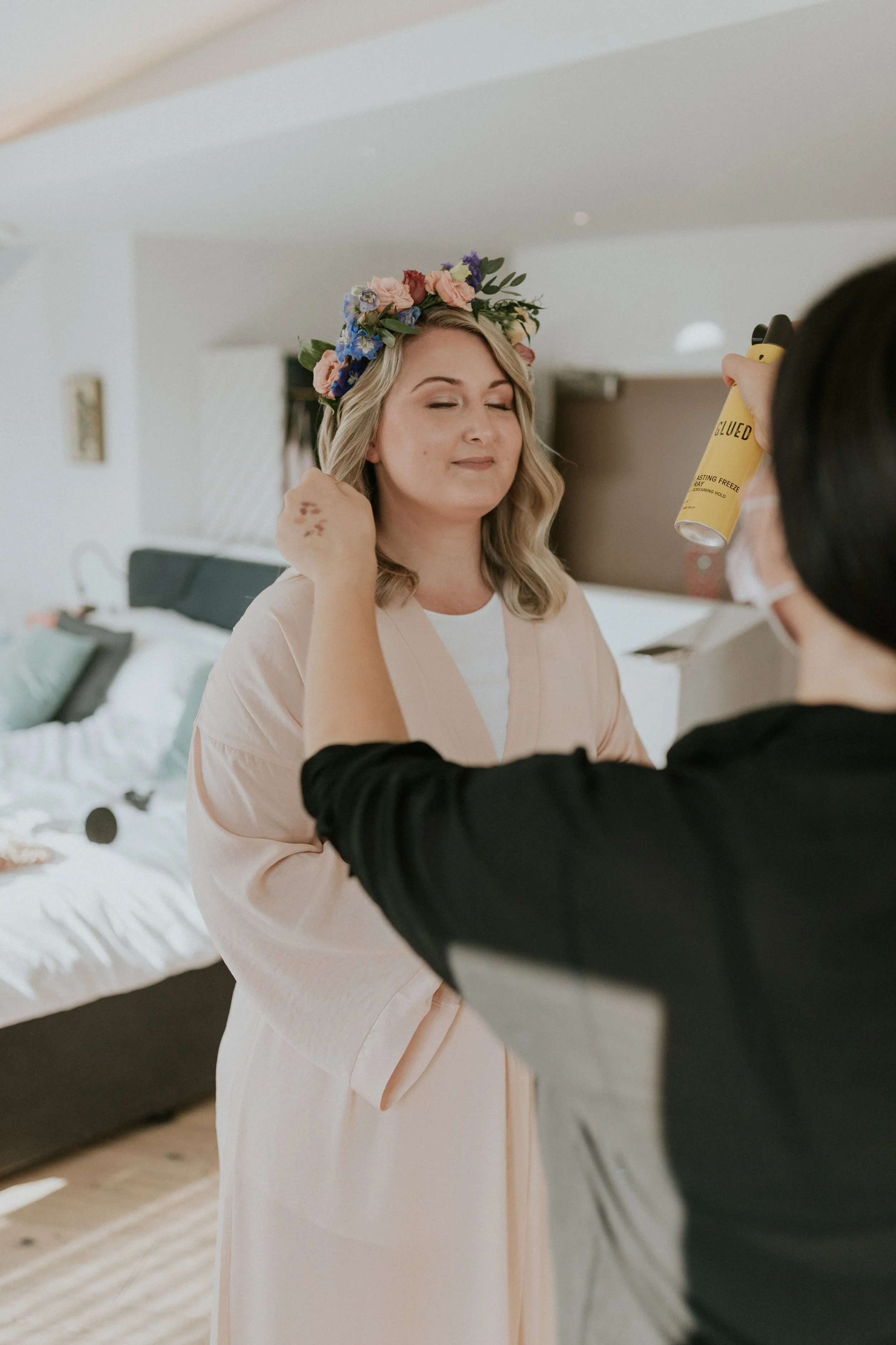 A woman with a floral crown and light pink robe is getting her makeup or hair done by a stylist in a bedroom with a bed in the background.