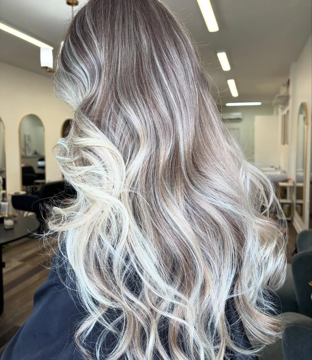 A woman with long, wavy hair styled in loose curls, featuring an ombre of dark roots transitioning to platinum blonde tips.