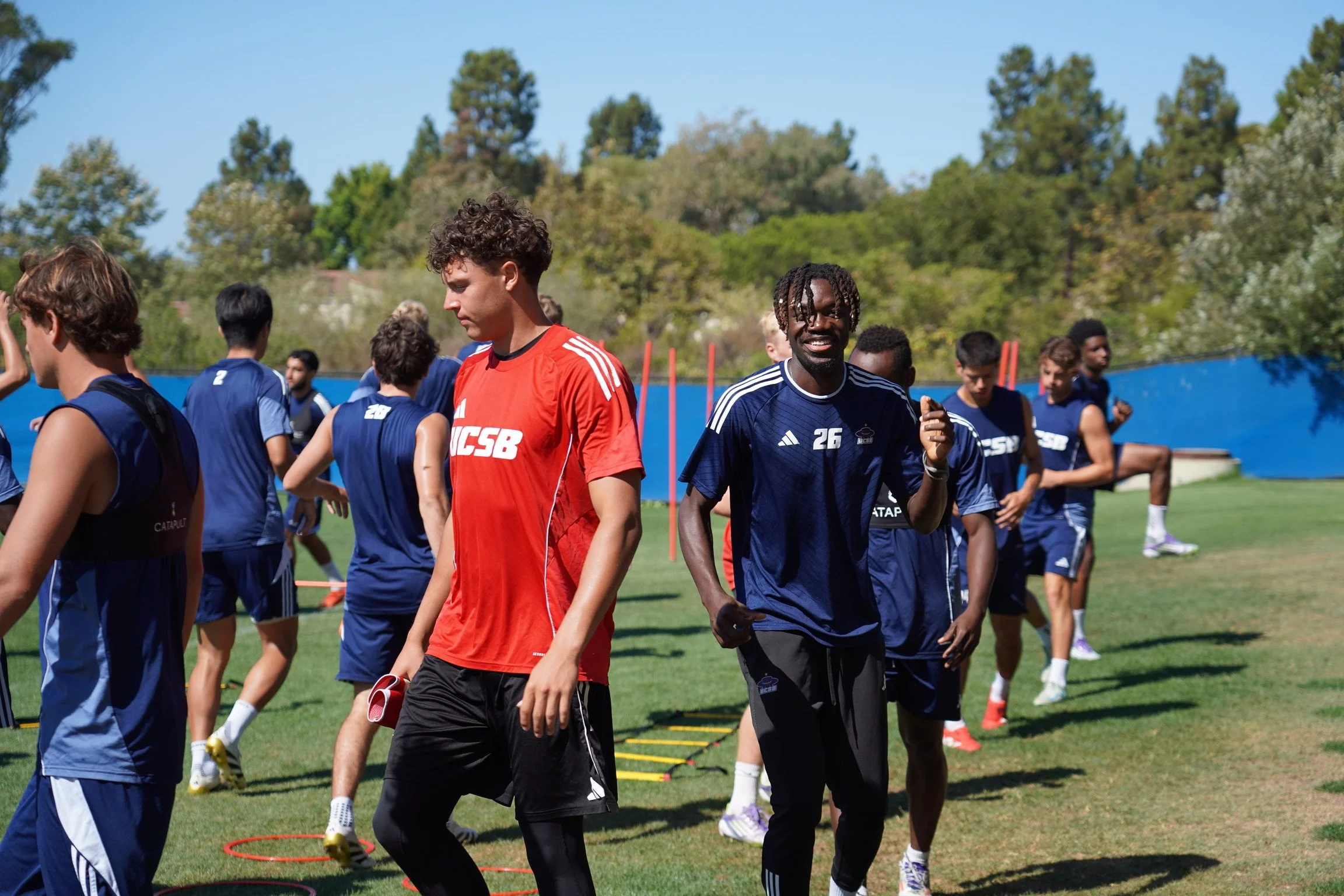 Soccer players in training on a grassy field, some walking and some stretching, with trees and a blue fence in the background.