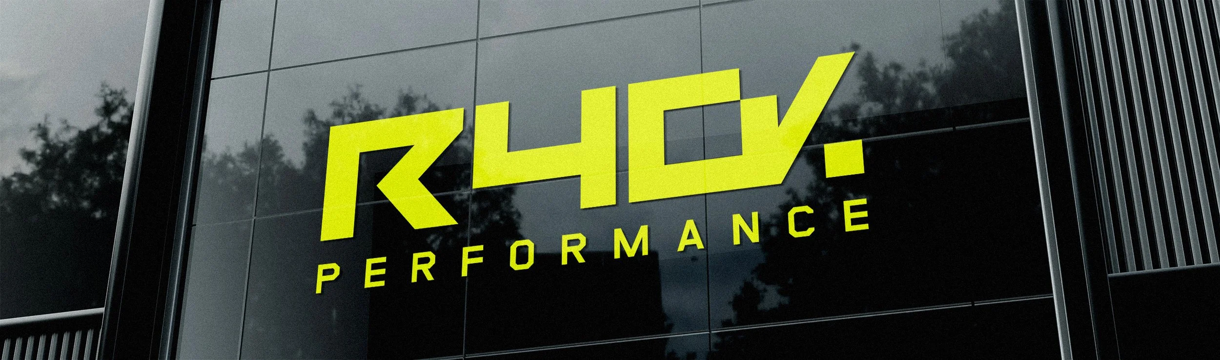 Sign on building with yellow text reading 'R4L0 PERFORMANCE' on a black reflective glass facade, with trees reflected in the glass.
