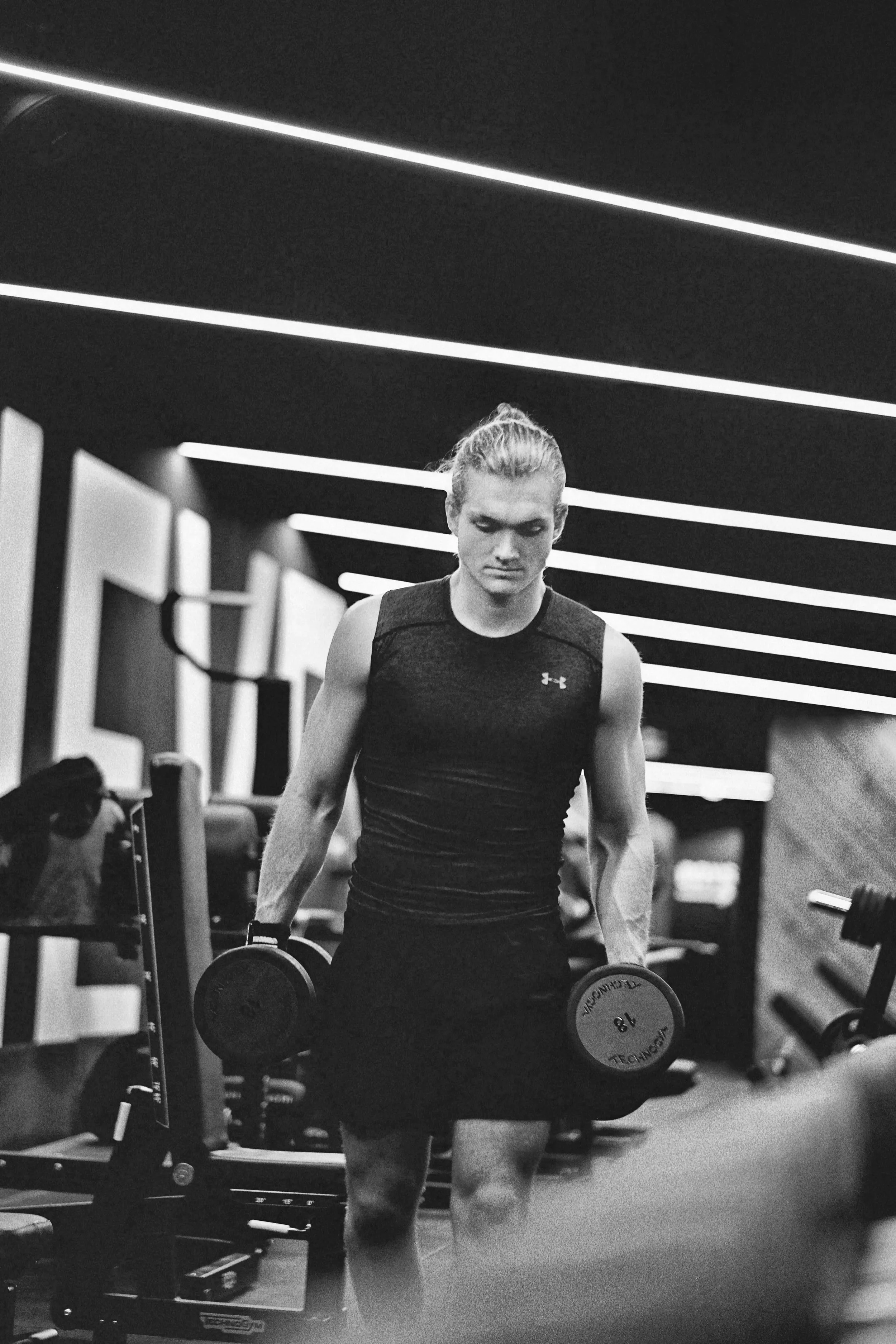 A woman lifting dumbbells in a gym, surrounded by workout equipment, with a modern ceiling design with illuminated lines.