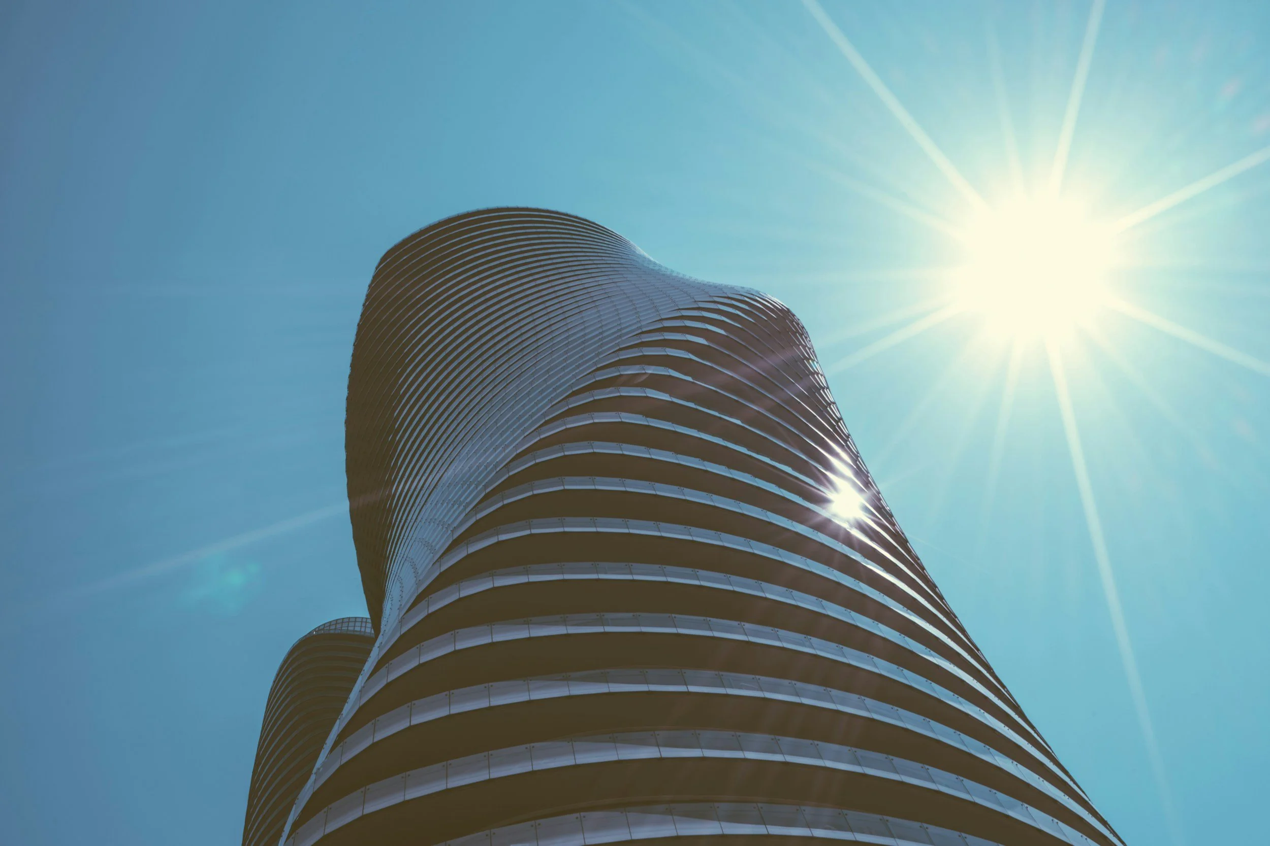 A modern high-rise building with curved architecture seen from below against a clear blue sky with the sun shining brightly.