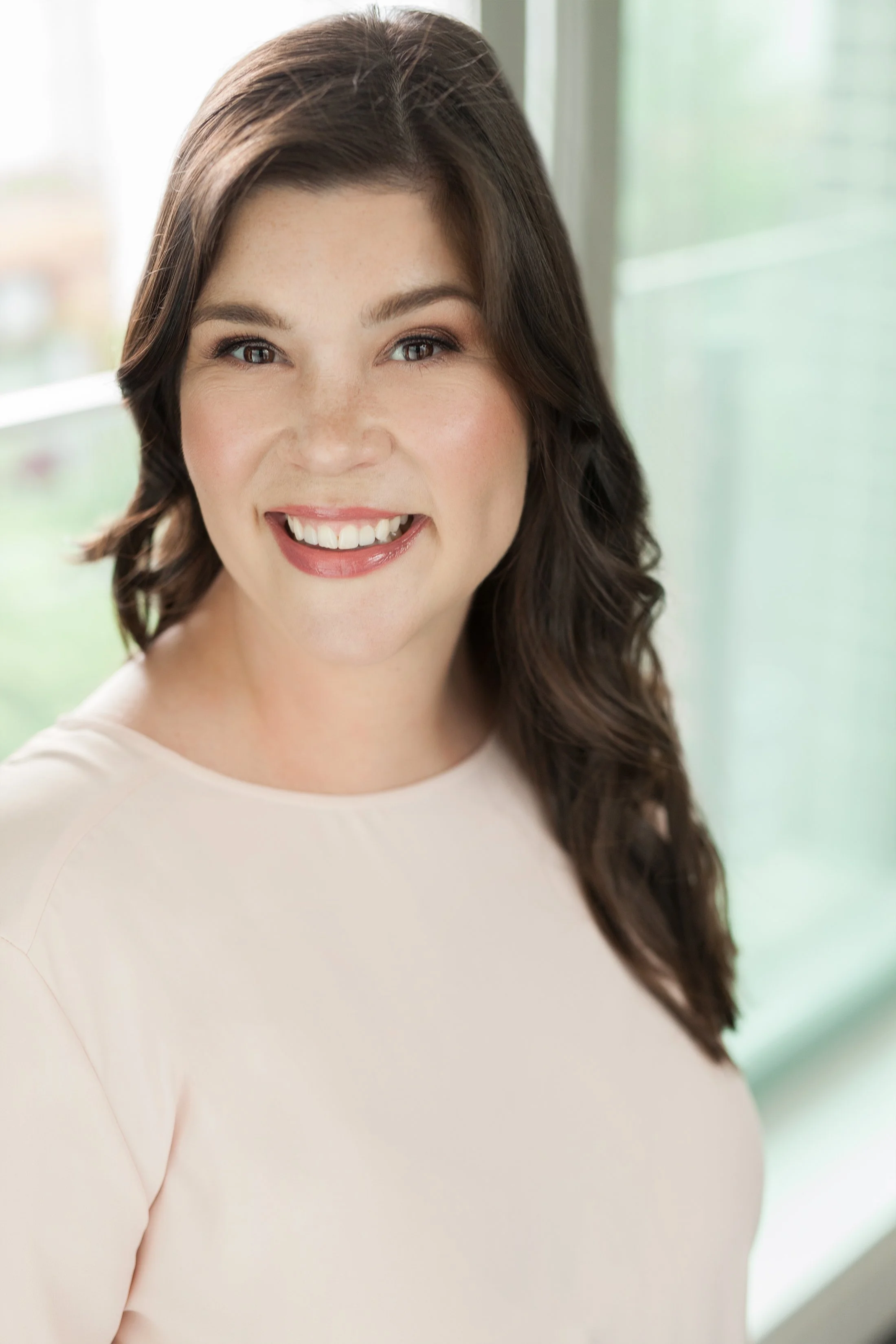Headshot of a smiling woman with medium-length dark hair, wearing a light-colored top, standing near a window with a blurred green outdoor background.