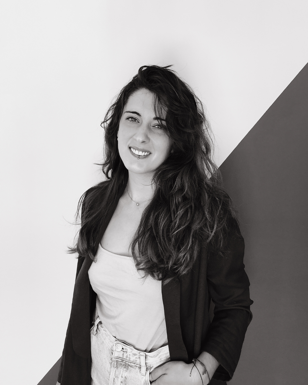 Retrato en blanco y negro de una mujer joven con cabello rizado, rubia oscura, sonriendo, usando una chaqueta y camiseta, con fondo dividido en blanco y gris.