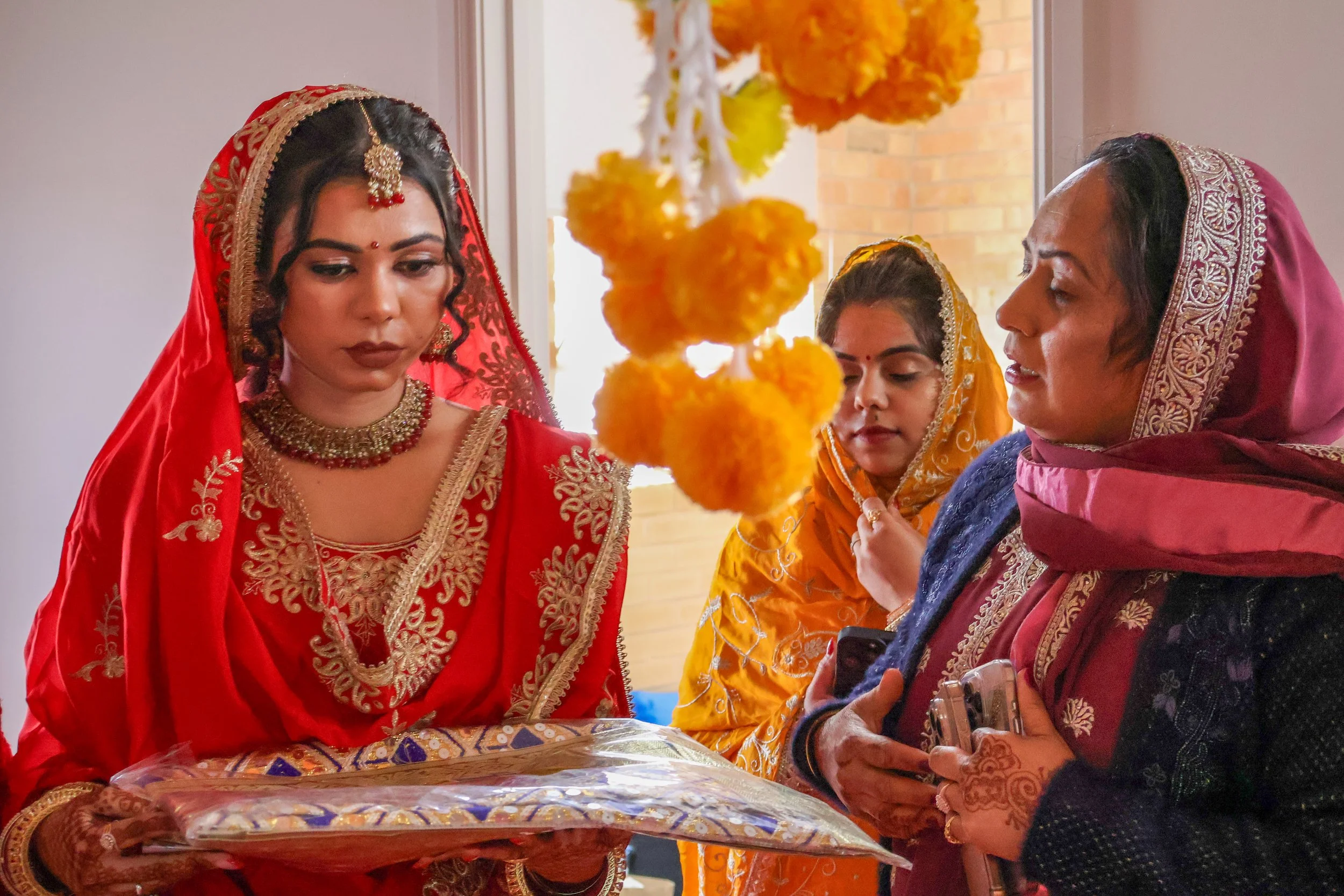 Women dressed in traditional Indian attire participating in a ceremony, with yellow marigold flowers hanging in the foreground.