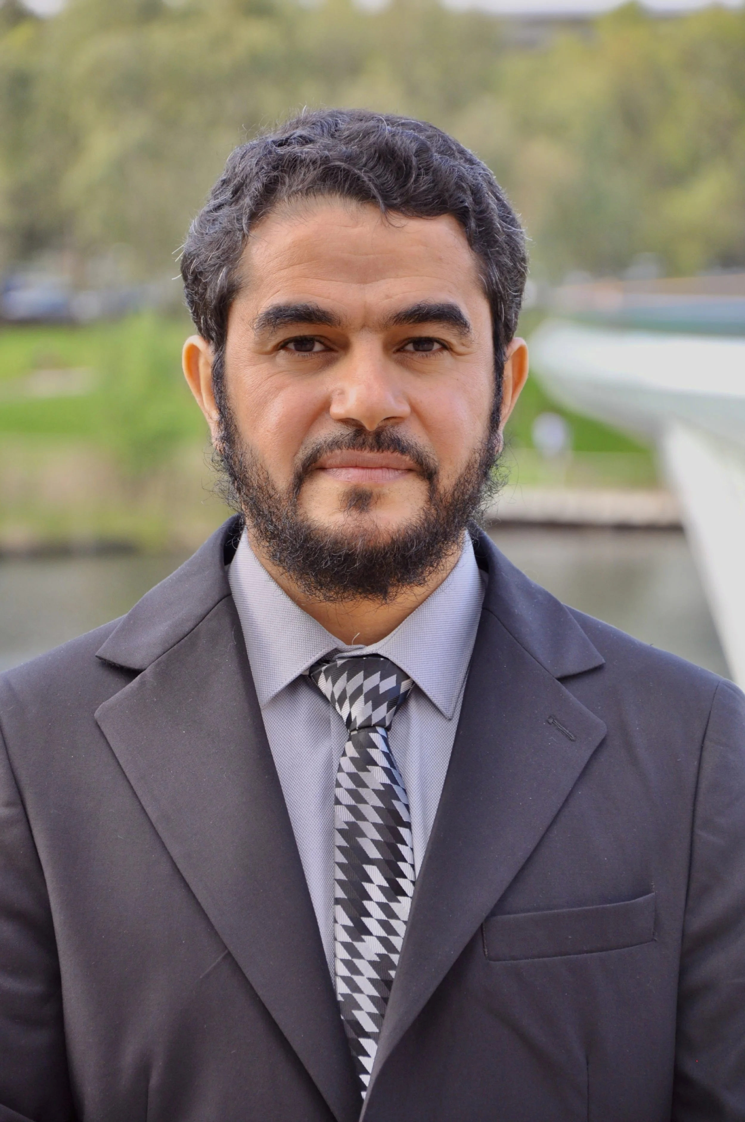 Portrait of a man in a business suit outdoors with a blurred background of trees and water.