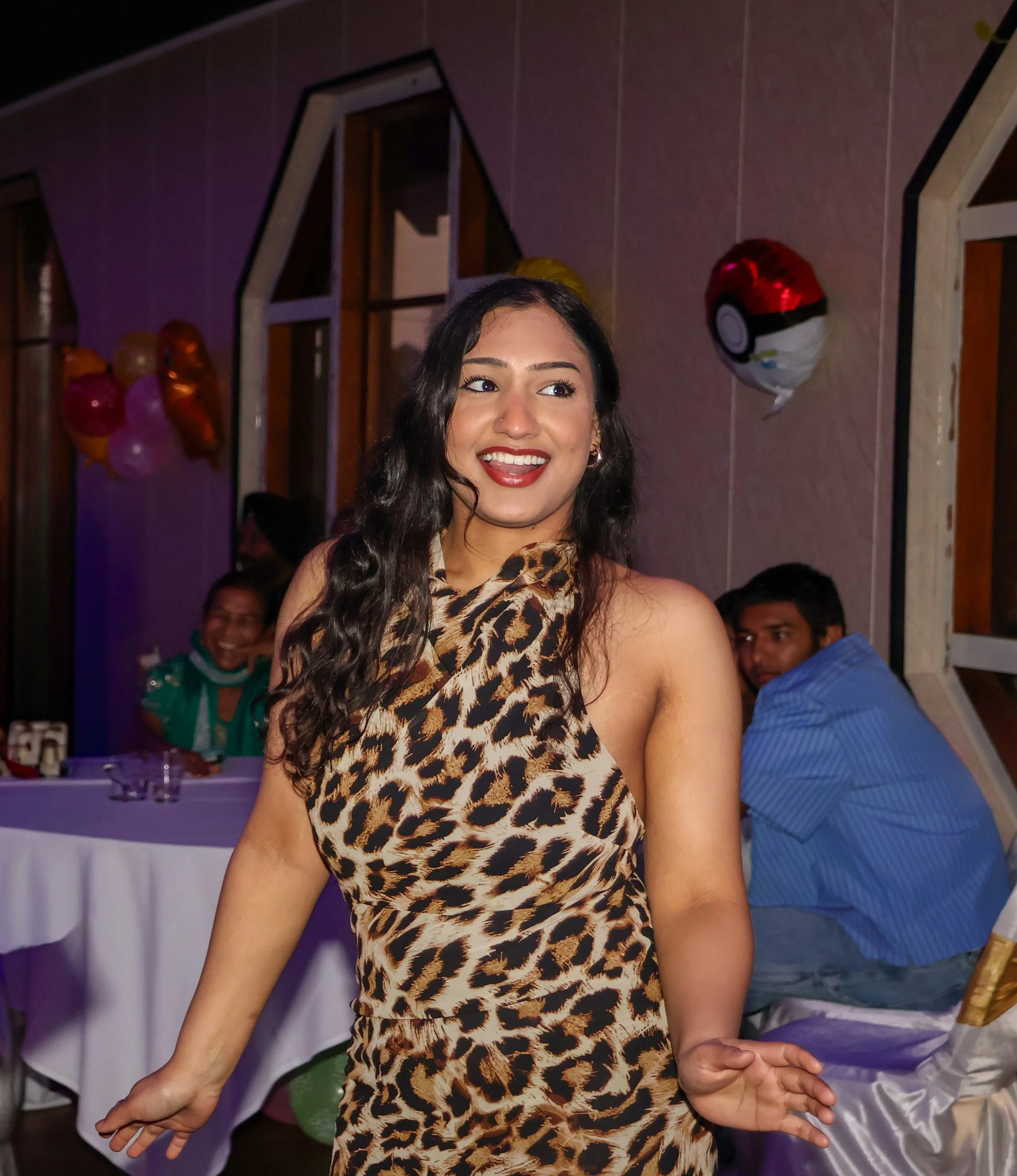 A woman with wavy dark hair and red lipstick wearing a leopard print dress, smiling and dancing at a party with balloons and guests in the background.