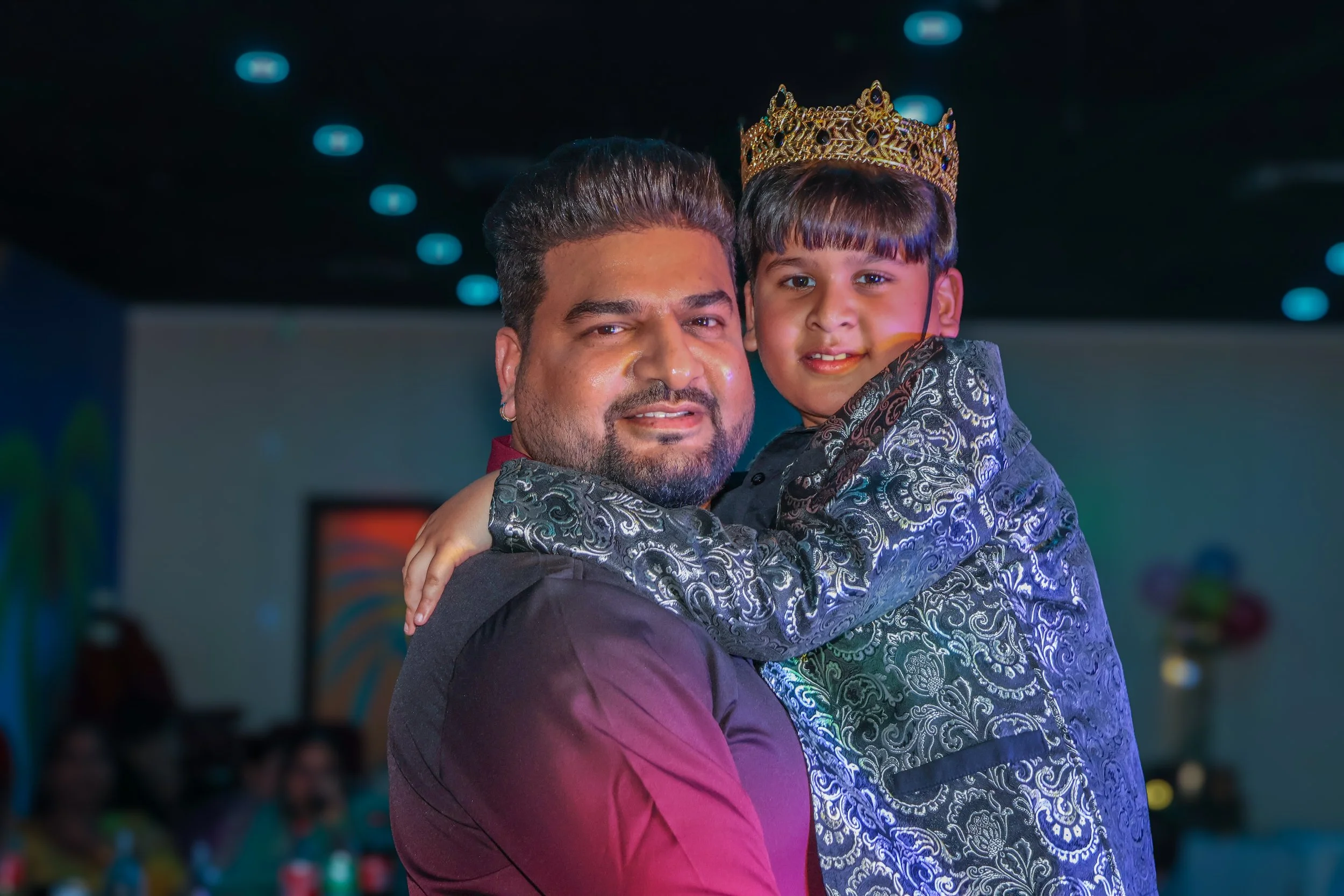 A man holding a young boy wearing a crown at a celebration or party indoors.