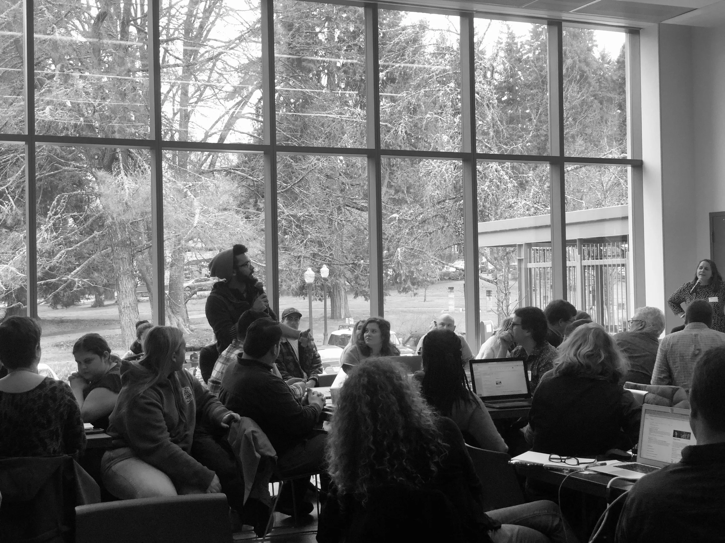 A group of people sitting in a conference room with large windows showing a snowy outdoor scene, some people are listening, while one man is speaking into a microphone.