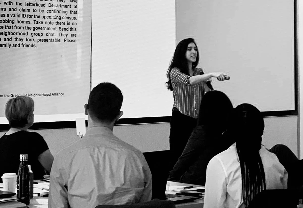 A woman giving a presentation in a conference room, pointing at a large screen with a remote control, while five people watch and listen.