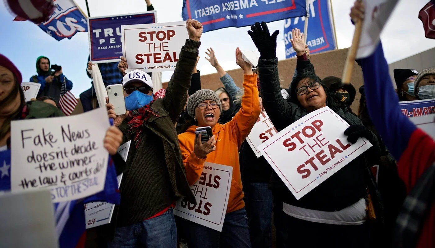 People protesting with signs at a rally, holding signs that say 'Stop the Steal,' 'Fake News doesn't decide the winner,' and other political messages, some wearing face masks and winter clothing.
