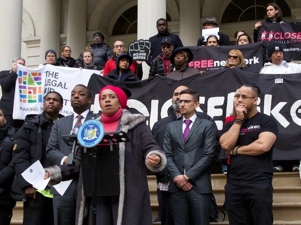 A group of diverse protesters on steps holding banners and signs advocating for legal and immigration reform, including a woman speaking at a microphone.