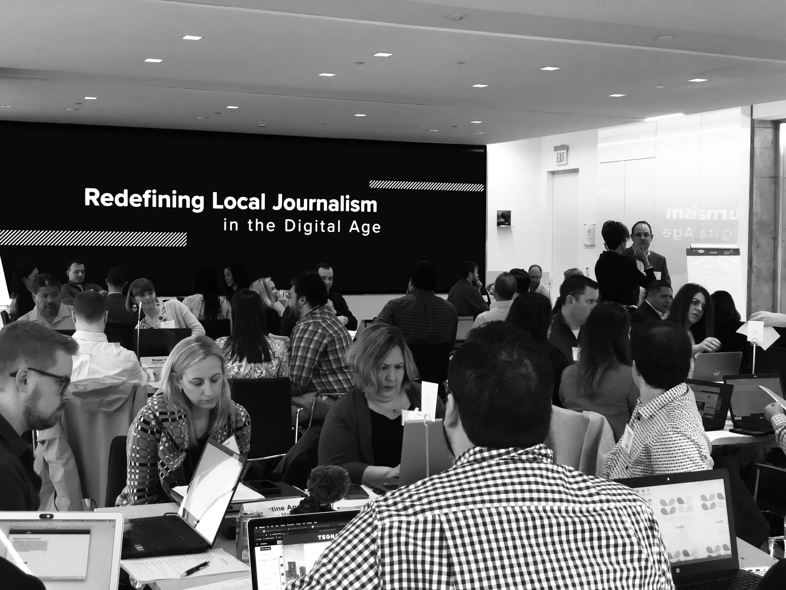 A conference room with many people sitting at tables, some working on laptops and others talking. There is a large screen on the wall displaying the text 'Redefining Local Journalism in the Digital Age'.