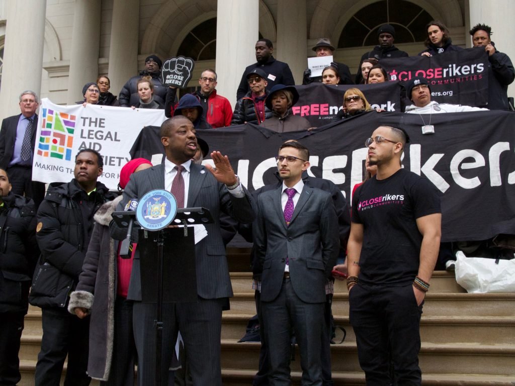 A diverse group of people gathered on the steps of a building holding banners and signs protesting. One man at a microphone appears to be speaking, with others listening or standing nearby. The signs include messages like 'The Legal Aid Society,' 'Close Rikers,' and 'Free New York,' indicating an advocacy event likely related to social justice or criminal justice reform.