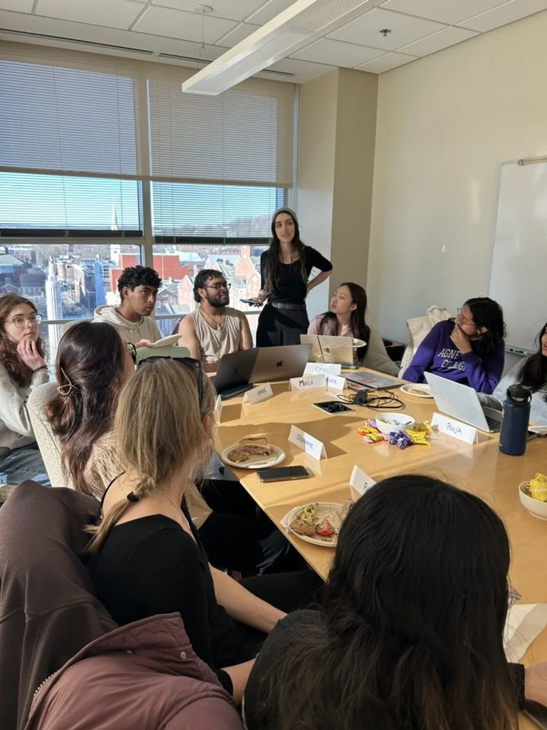 A group of people sitting around a conference table in a meeting room with large windows and a city view, listening to a woman standing and speaking.