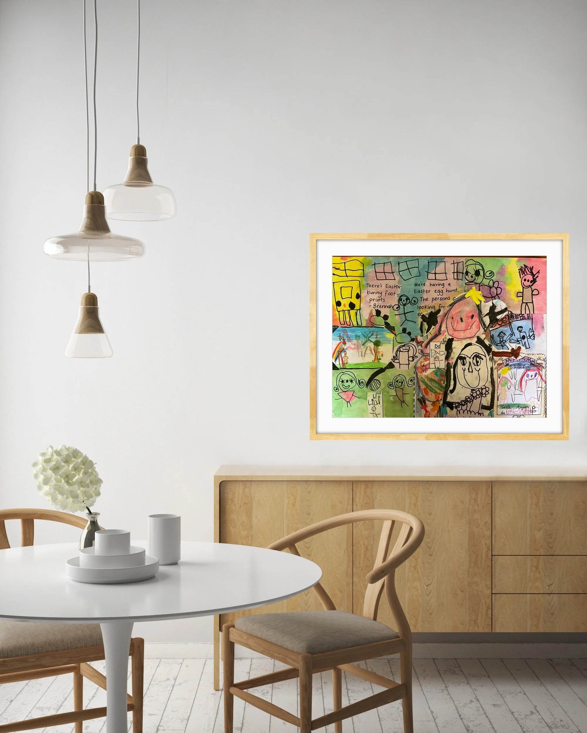 Children's colorful drawing with animals, houses, and text, framed and hanging on a white wall in a minimalist dining room with wooden furniture.