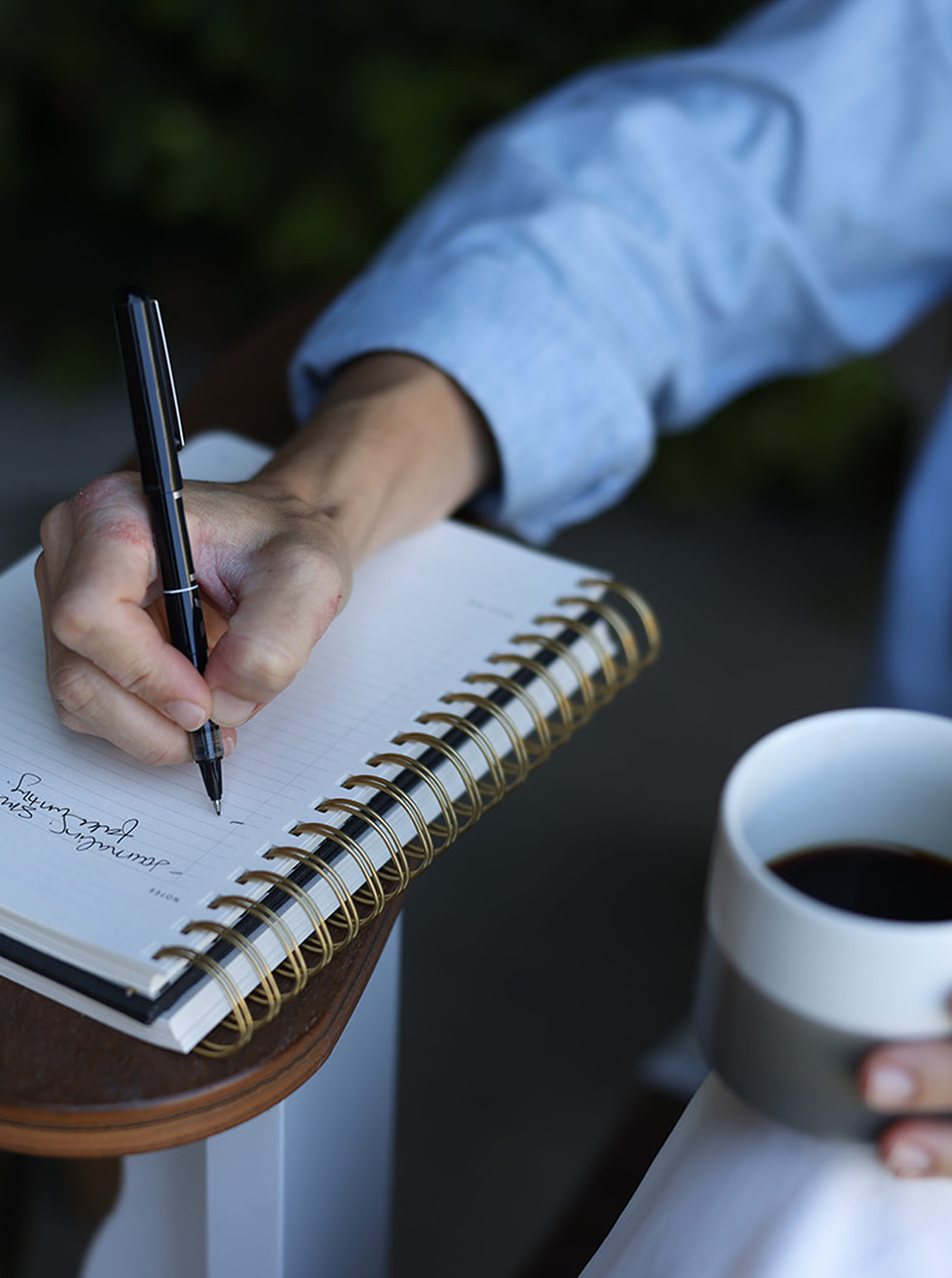 Person in a light blue shirt writing in a spiral notebook with a black pen, holding a cup of coffee.