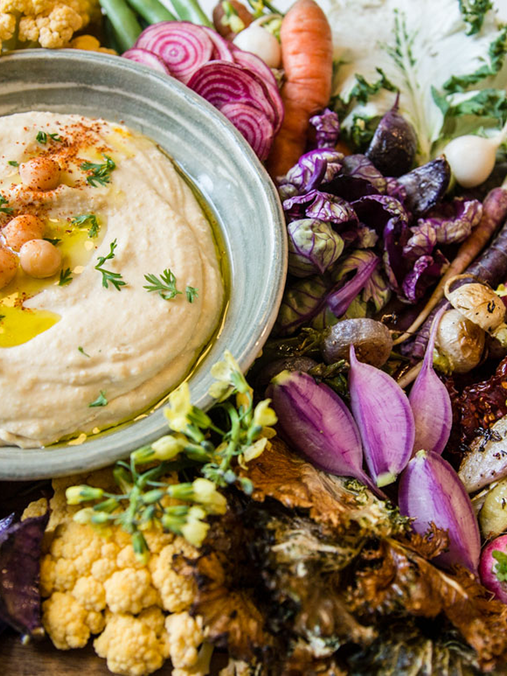A bowl of hummus garnished with chickpeas, parsley, and olive oil surrounded by fresh vegetables including cauliflower, purple onions, carrots, radishes, purple cabbage, and garlic.