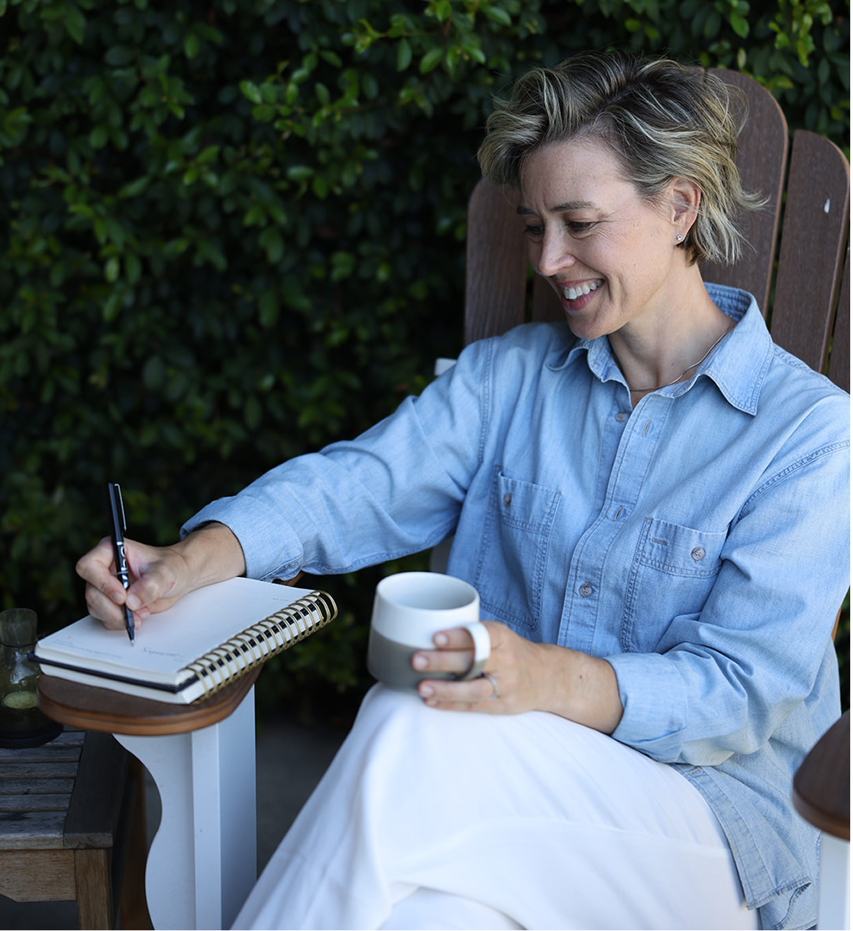 A woman with short, gray hair, wearing a light blue shirt and white pants, smiling while writing in a spiral notebook with a pen, sitting outdoors in a wooden chair holding a mug.