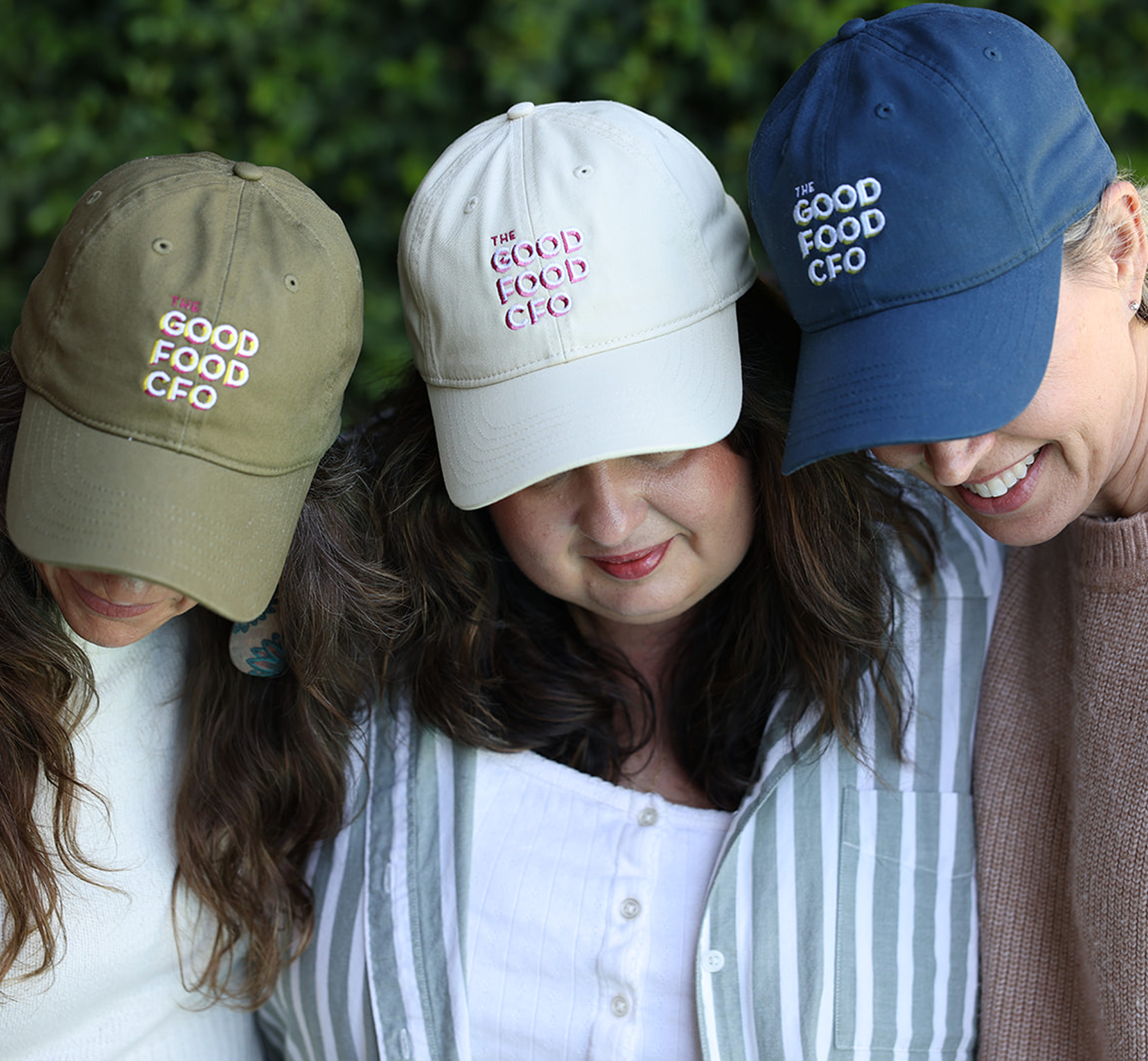 Three women wearing baseball caps with the words 'The Good Food CFO' embroidered on them, standing close together outdoors.