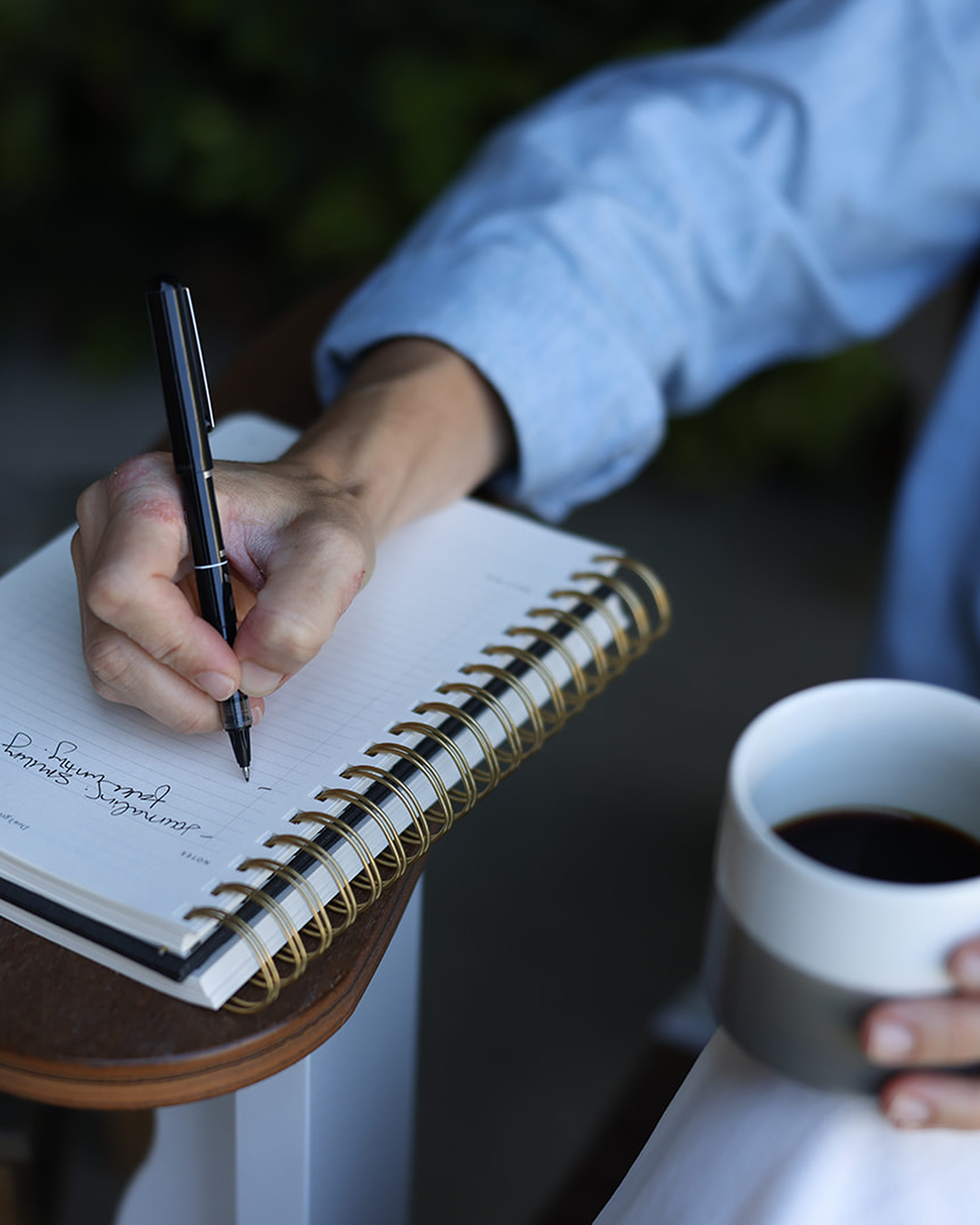 A person wearing a light blue shirt writing in a spiral-bound notebook with a black pen, holding a cup of coffee in their other hand, sitting at a wooden table.