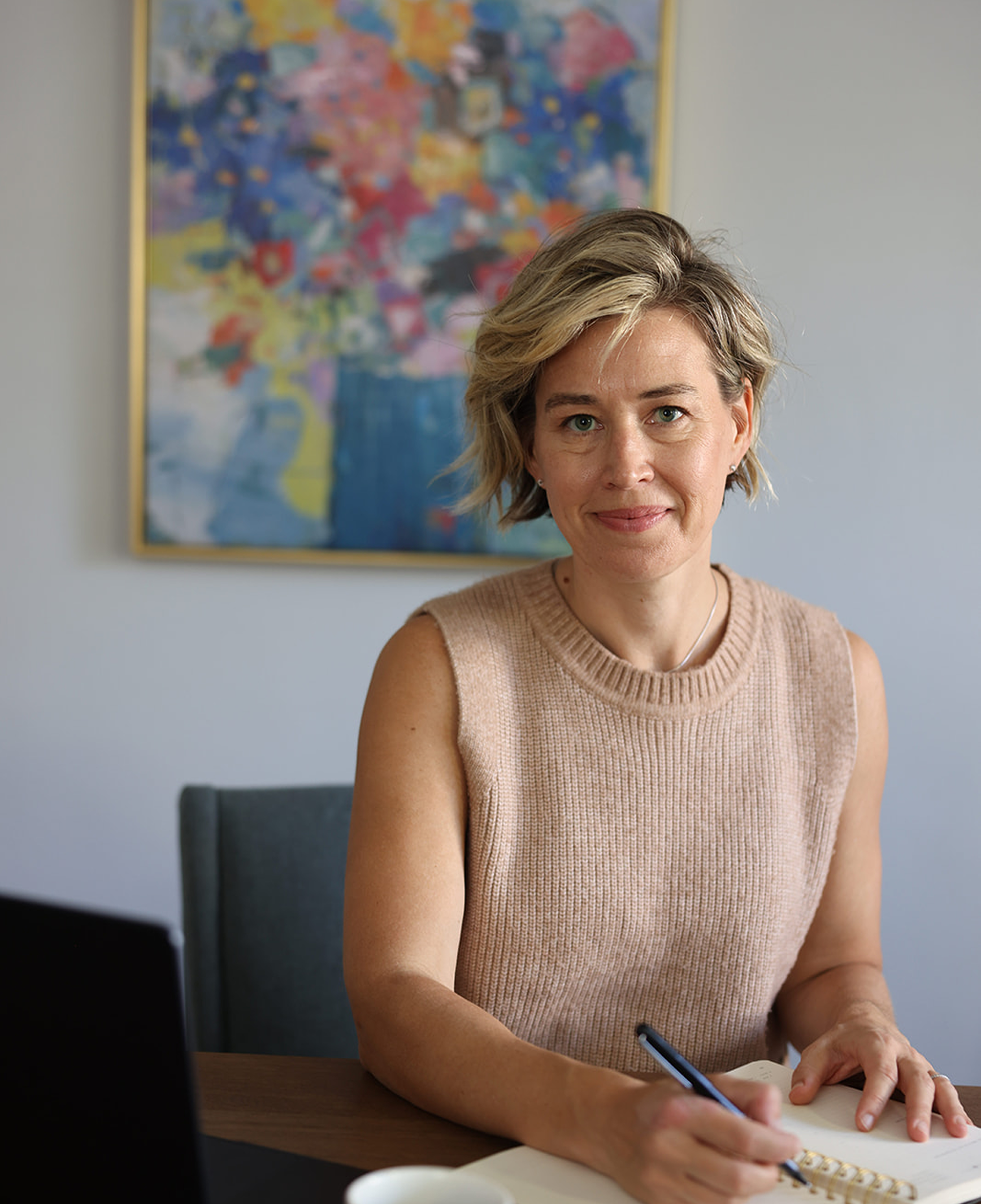 A woman with short blonde hair wearing a sleeveless beige knit top sitting at a desk, writing in a notebook with a pen, with a colorful abstract painting on the wall behind her.