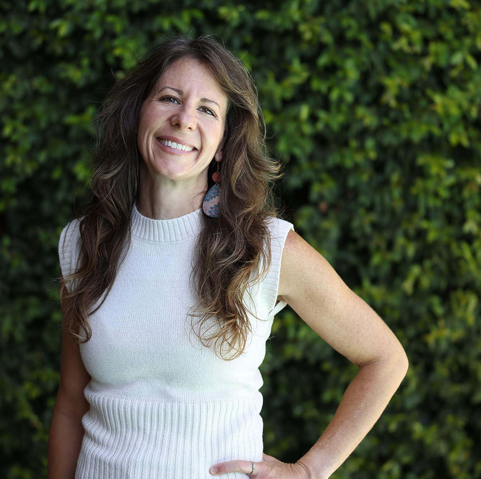 A woman with long wavy brown hair smiling outdoors against a green leafy background, wearing a sleeveless white knit top and colorful earrings.