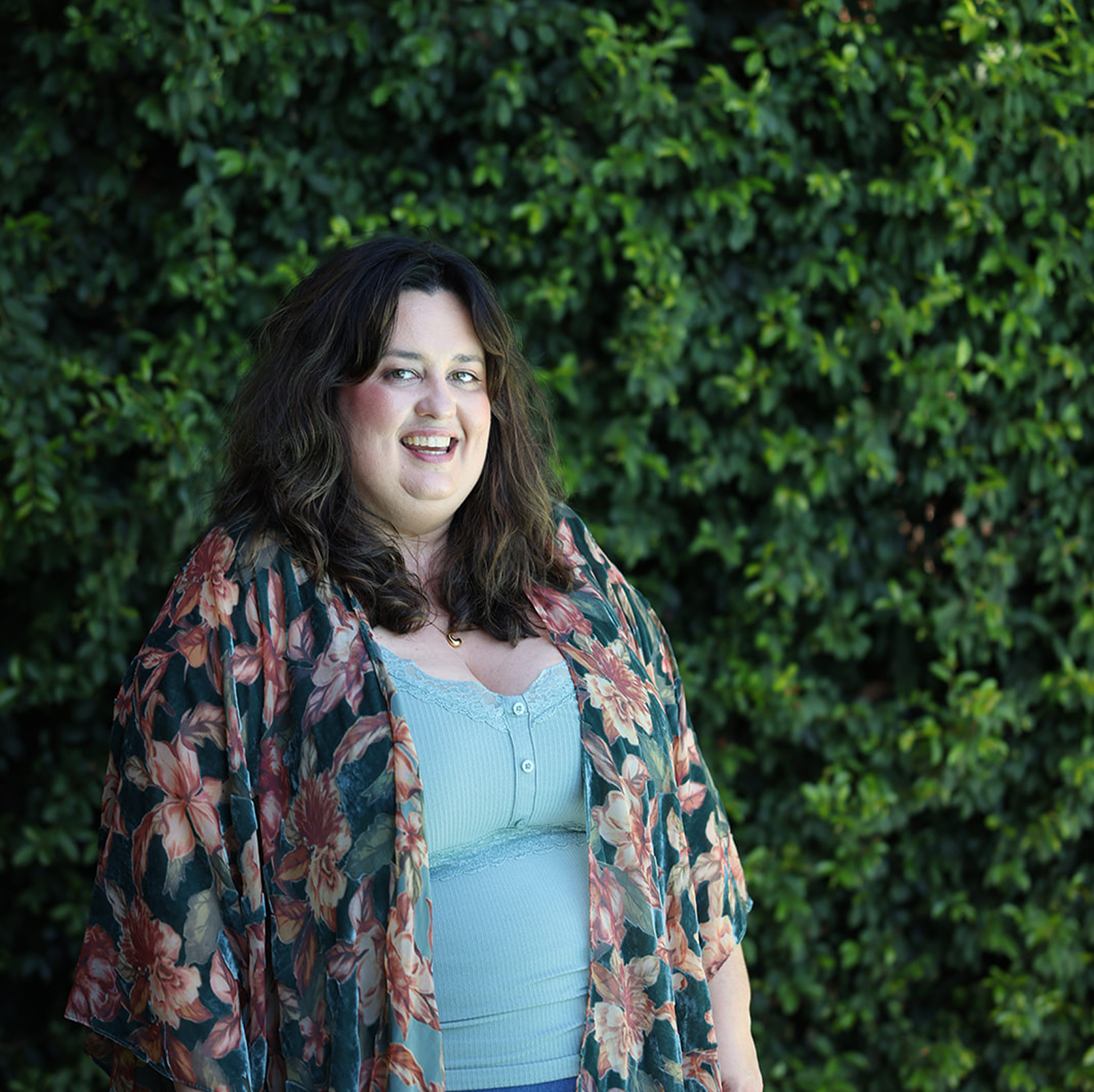 Woman with long dark hair wearing a floral jacket and light blue shirt standing in front of greenery.
