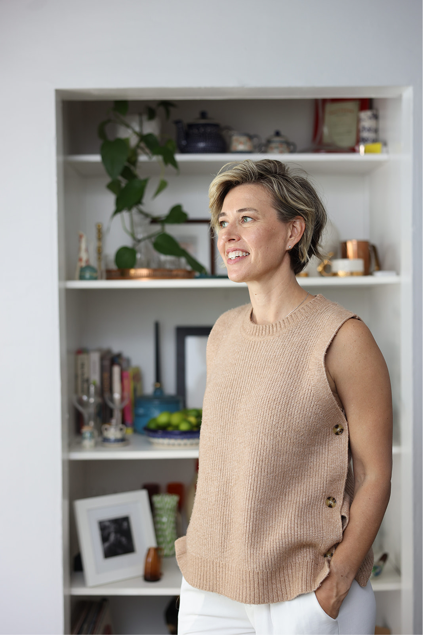 A woman with short, blonde hair smiling and looking to her left, standing in front of a white bookshelf filled with decorative items, plants, books, and framed photos.