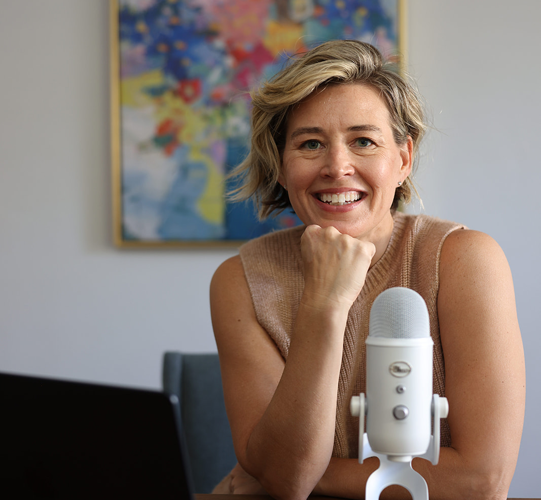 A woman with short blonde hair smiling at a microphone during a podcast or recording session, with a colorful world map hanging on the wall behind her.