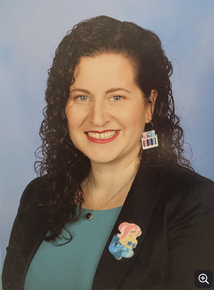 A woman with curly dark hair smiling at the camera, wearing a blue top and black blazer, with a mermaid pin and a colorful earring featuring bars.