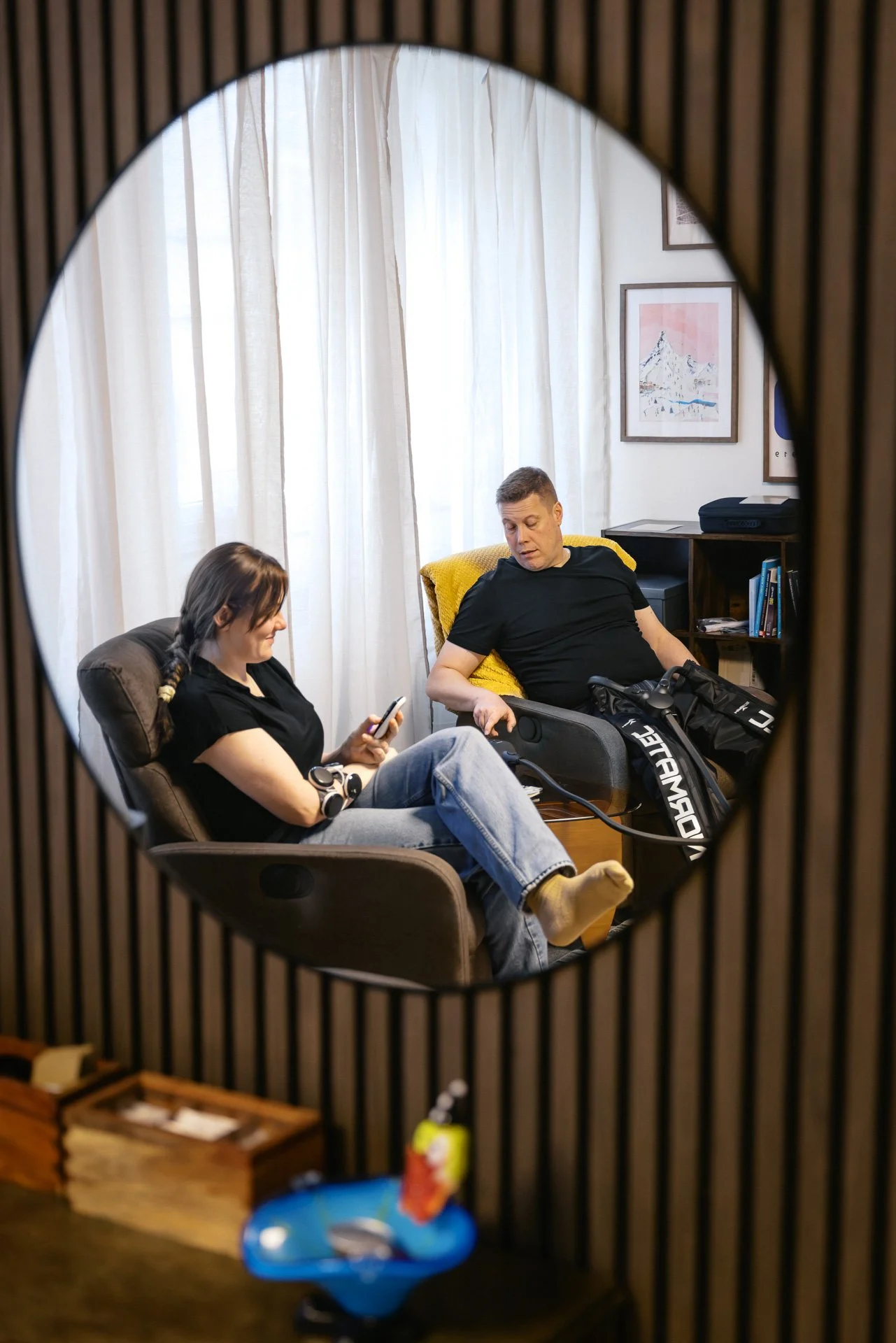 Två personer sitter i ett rum, en kvinna som tittar på sin mobiltelefon och en man som tittar på henne. De är omgivna av möbler och konst på väggarna.