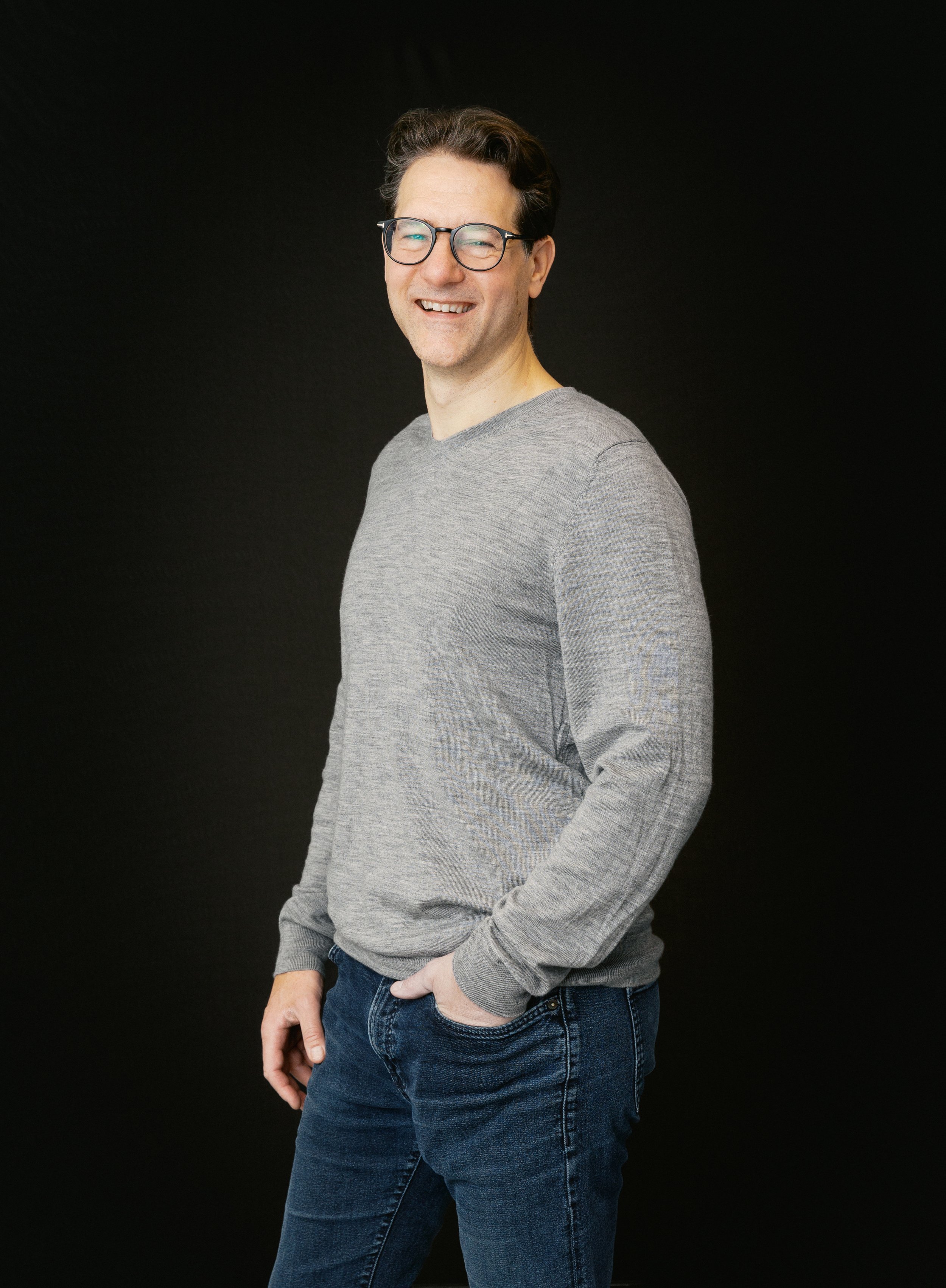 En ung man med glasögon, grå t-shirt och jeans som ler mot kameran mot en svart bakgrund.