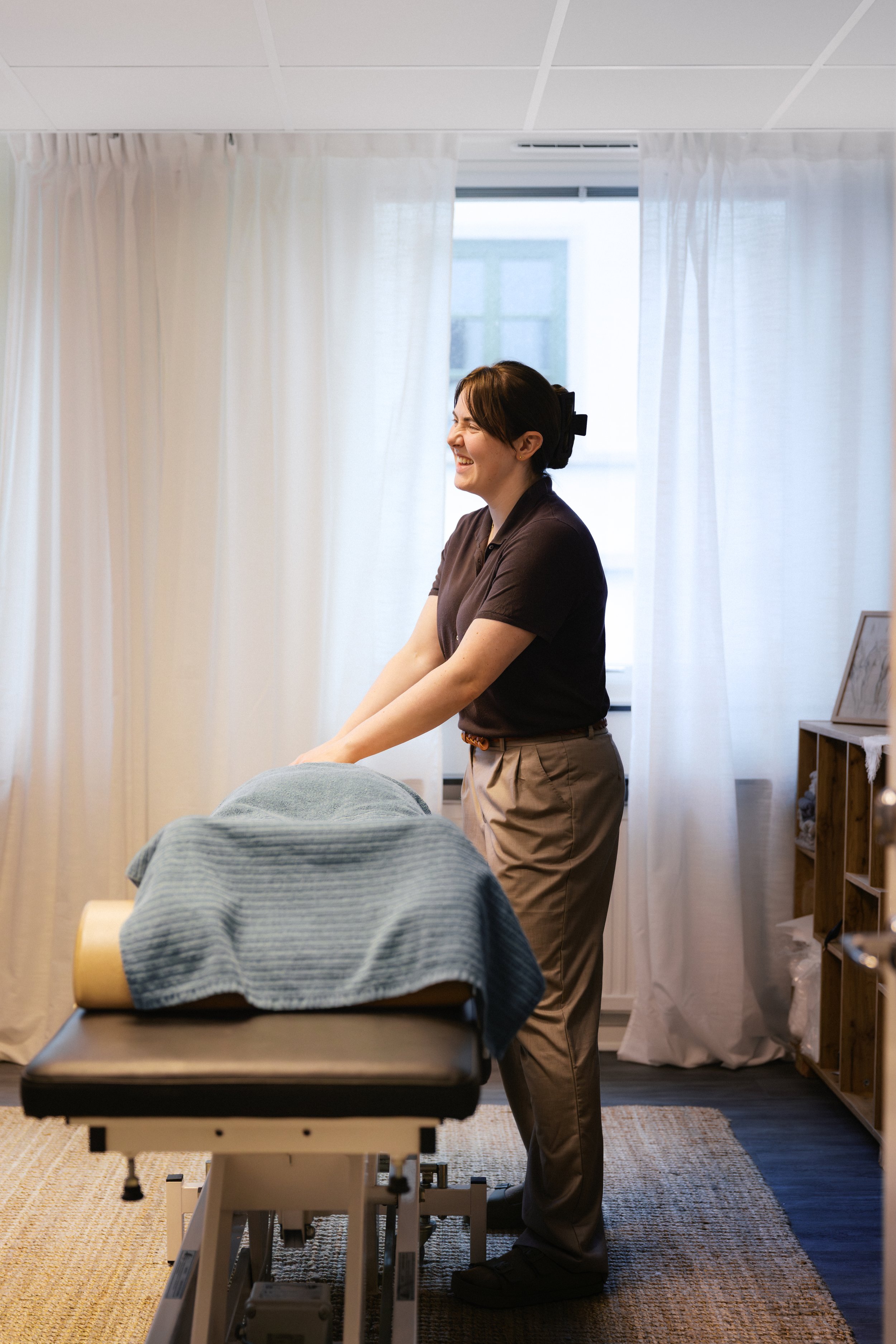 En kvinna i arbetskläder som ler medan hon står bredvid en massagebänk i ett rum med vita väggar och stora fönster med vita gardiner.