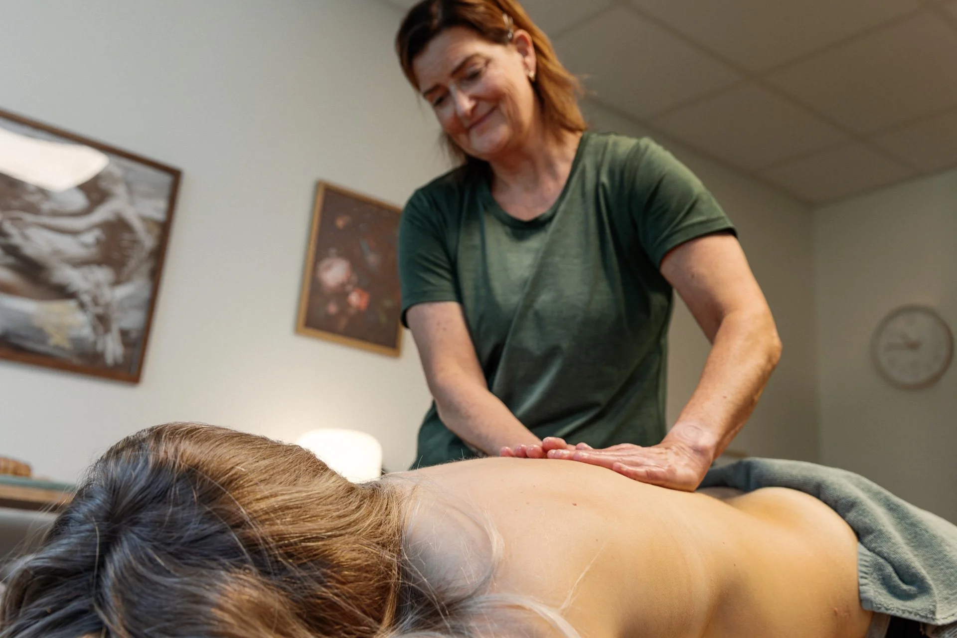 Kvinna som ger en massage till en person som ligger på mage i ett rum med tavla och väggklocka.