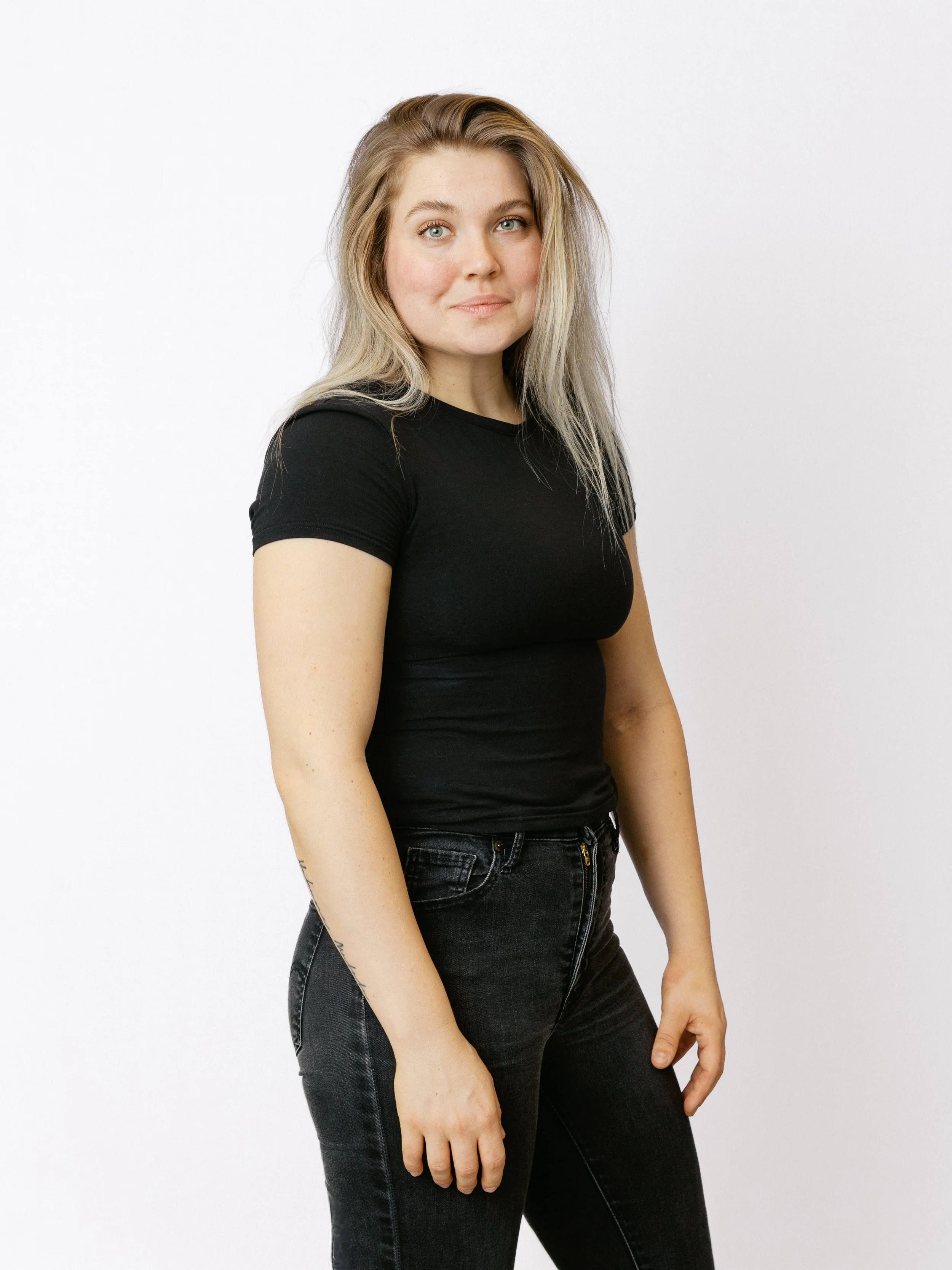 Ung kvinna med blont, vågigt hår, i svart T-shirt och jeans, står framför en vit vägg.