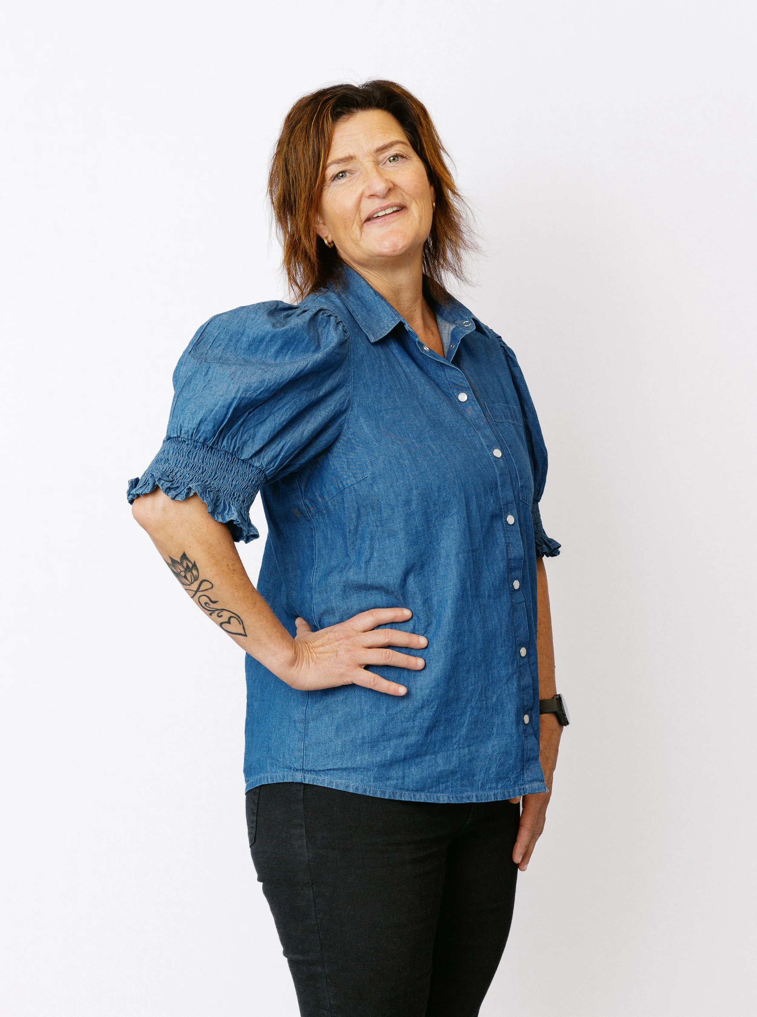 En kvinna med brunt lockigt hår, iklädd en blå denimklänning och svarta byxor, står mot en vit bakgrund.